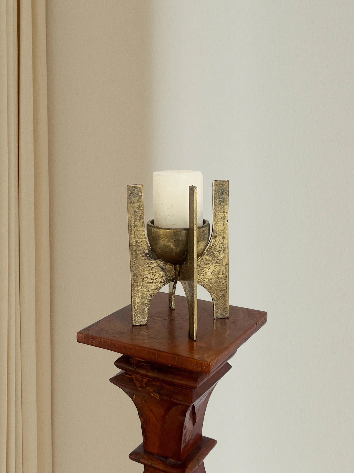 A white candle sits in Porter Studio's Brutalist Brass Candle Holder on a carved wooden pedestal, set against a backdrop of a light-colored wall and curtain, creating an elegant atmosphere with textured surfaces and timeless allure.