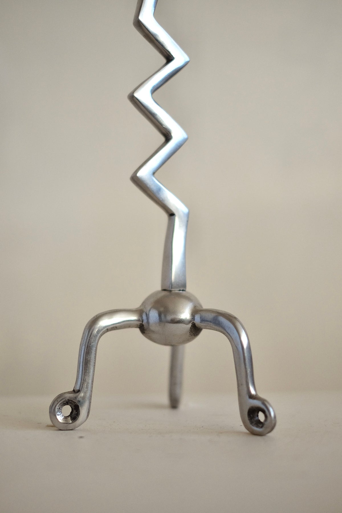 A silver item from the Set of Zigzag Candleholders by septembre studios stands upright with a round base and three curved legs ending in small circular loops, reminiscent of rare candle holders. The background's neutral tone accentuates the unique craftsmanship of this extraordinary find.