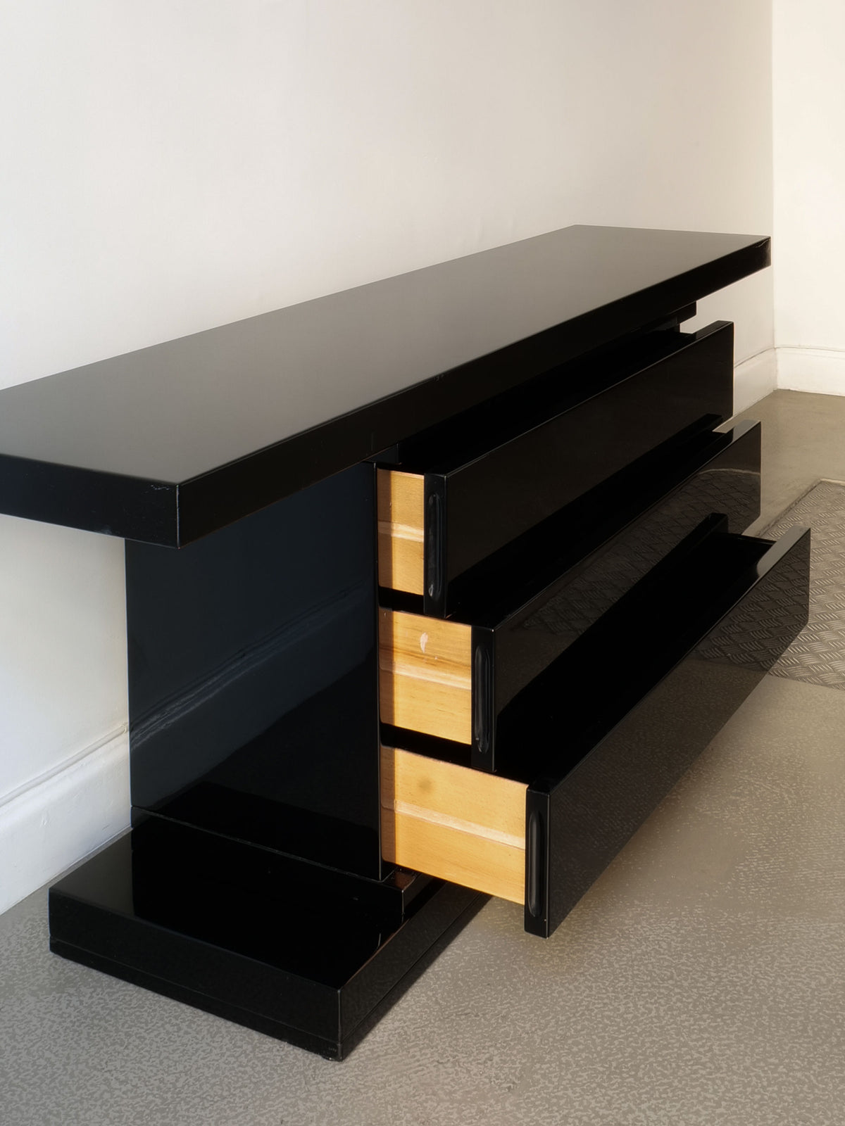 The High Gloss Console Table 1970s by Collection apart showcases a modern, minimalist Italian design with a sleek black exterior and high gloss finish. It features three open drawers that reveal light wood interiors, positioned against a white wall on a textured gray floor.
