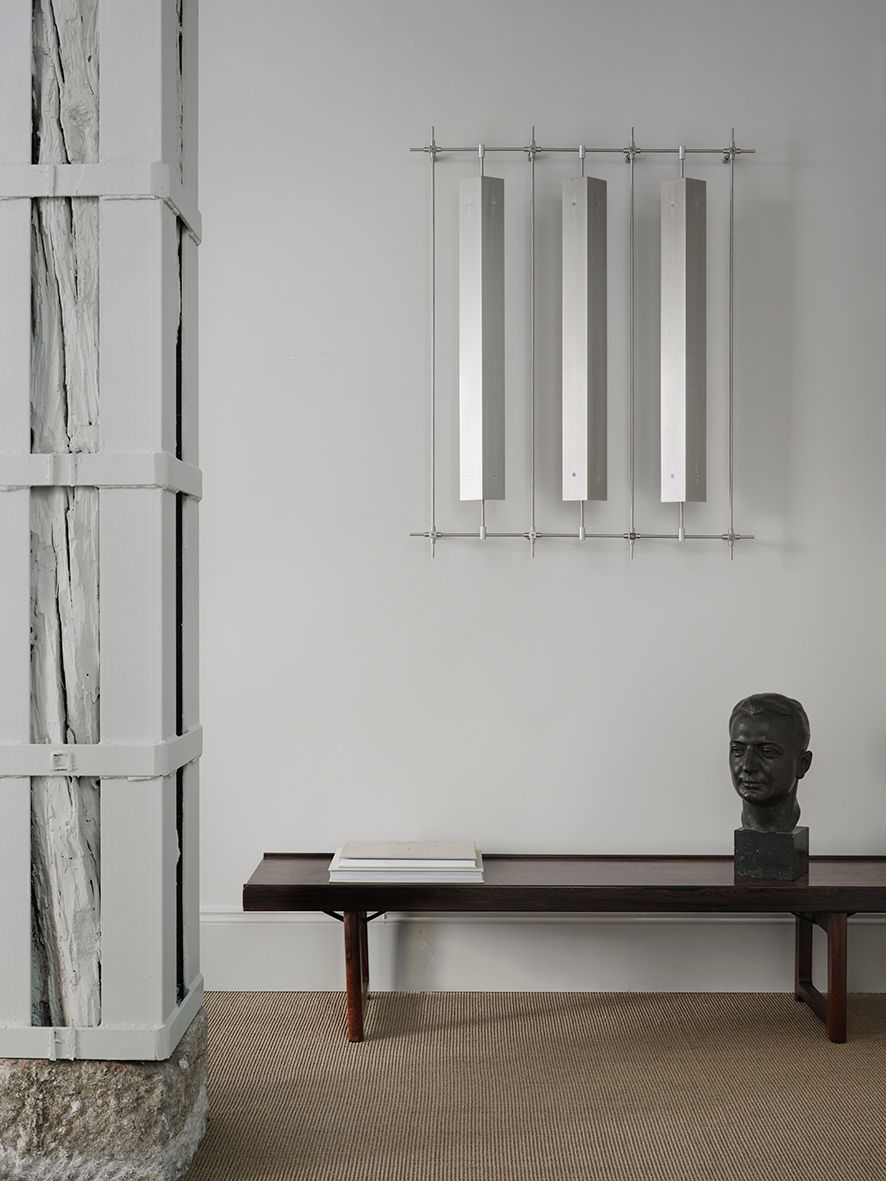 A minimalist interior features a dark wooden bench with a book and the black "Testa Virile" Sculpture 1930s by FEBRERO SHOP, set against a gray wall with three vertical metallic panels. A structural column with white and gray 1930s-style accents stands to the left.