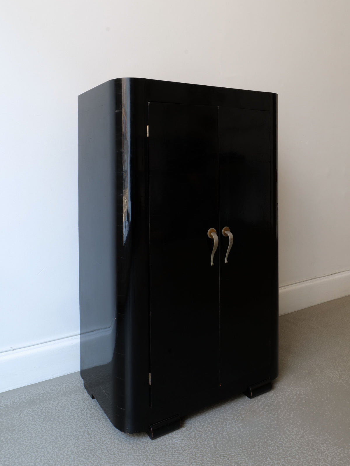 The French Deco Cabinet 1930s by Collection Apart is a sleek, black lacquer cabinet with curved edges and two vertical doors with silver handles, evoking a French 1930s design, against a light wall on a gray floor.