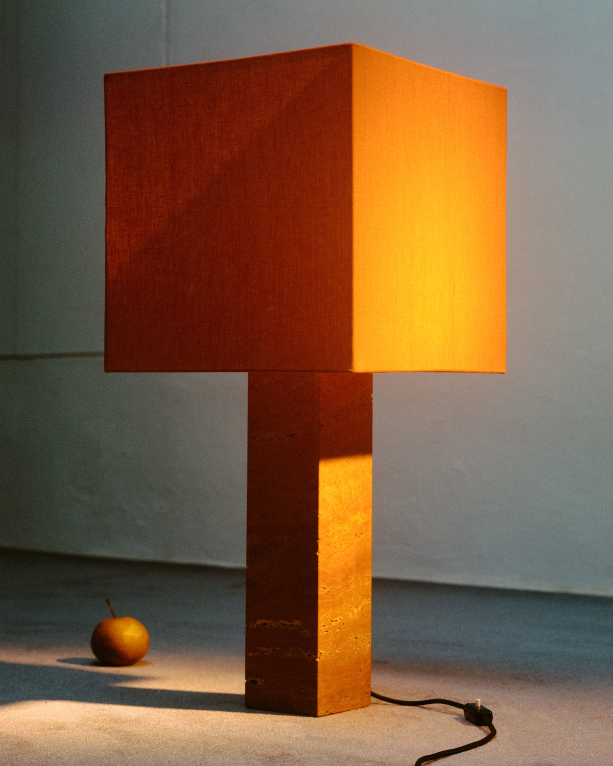A square-shaped lamp with a warm orange shade and a matching rectangular base stands on a minimalist floor, emphasizing modernism. Made from pure linen lampshade and other natural materials, its cord is visible near the base. A single pear lies next to the Roma I Squared Red Travertine by Nassi Lamps, casting a shadow in the ambient light.