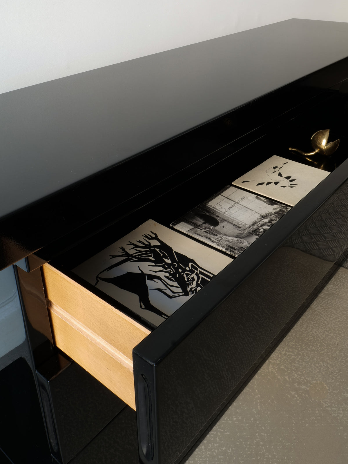 A sleek, black High Gloss Console Table from the 1970s by Collection apart showcases two black-and-white art prints and a brass-colored spoon in its open drawer. The custom-built piece stands against a light-colored wall, with the polished concrete floor enhancing its elegance.
