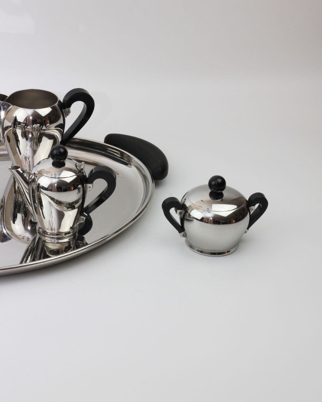 Introducing the "Bombè" Alessi Tea/Coffee Set from Boga Avante Shop, an exclusive collection showcasing mid-20th-century Italian craftsmanship. The set features a stainless steel teapot, sugar bowl, and creamer with black handles, all presented on a coordinated tray. Additionally, a small lidded sugar bowl by Alfra Alessi is positioned to the right of the tray against a plain white background.