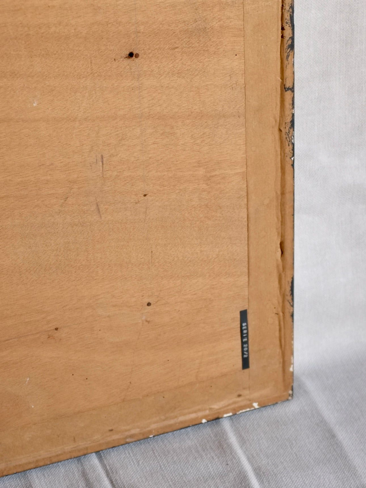 A close-up view of the back of a wooden picture frame with a small label on the lower right corner. The frame, likely enclosing a Vintage Genre Oil Painting with Market Scenes by renowned Spanish artist under the brand Veter Vintage, is resting on a light-colored surface.
