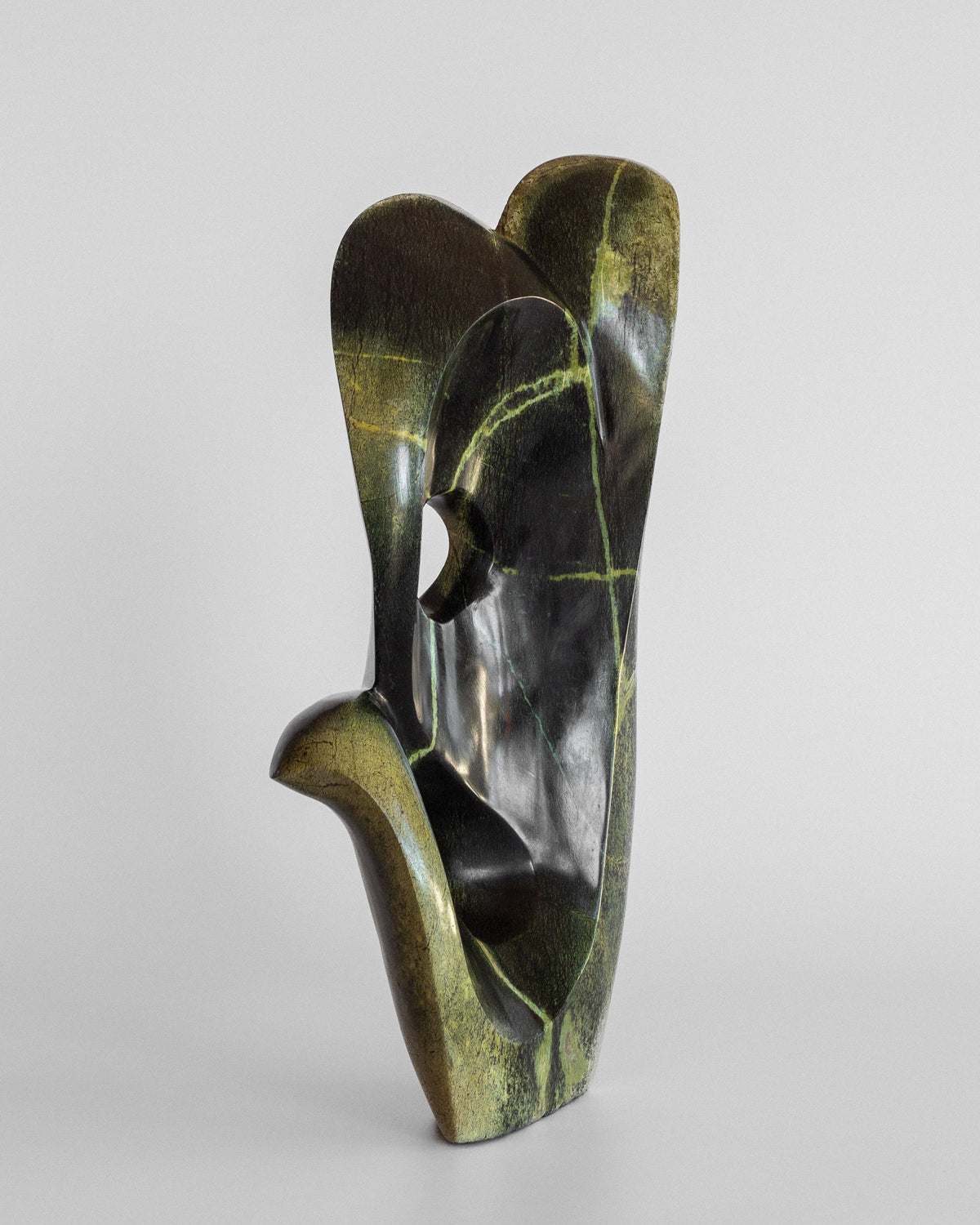 Abstract Stone Sculpture II" by Spigel is a Zimbabwean piece crafted from smooth leopard stone, showcasing abstract, organic shapes with curves and a small circular hole. It features earthy green and black hues with subtle yellow and white veins against a plain background.
