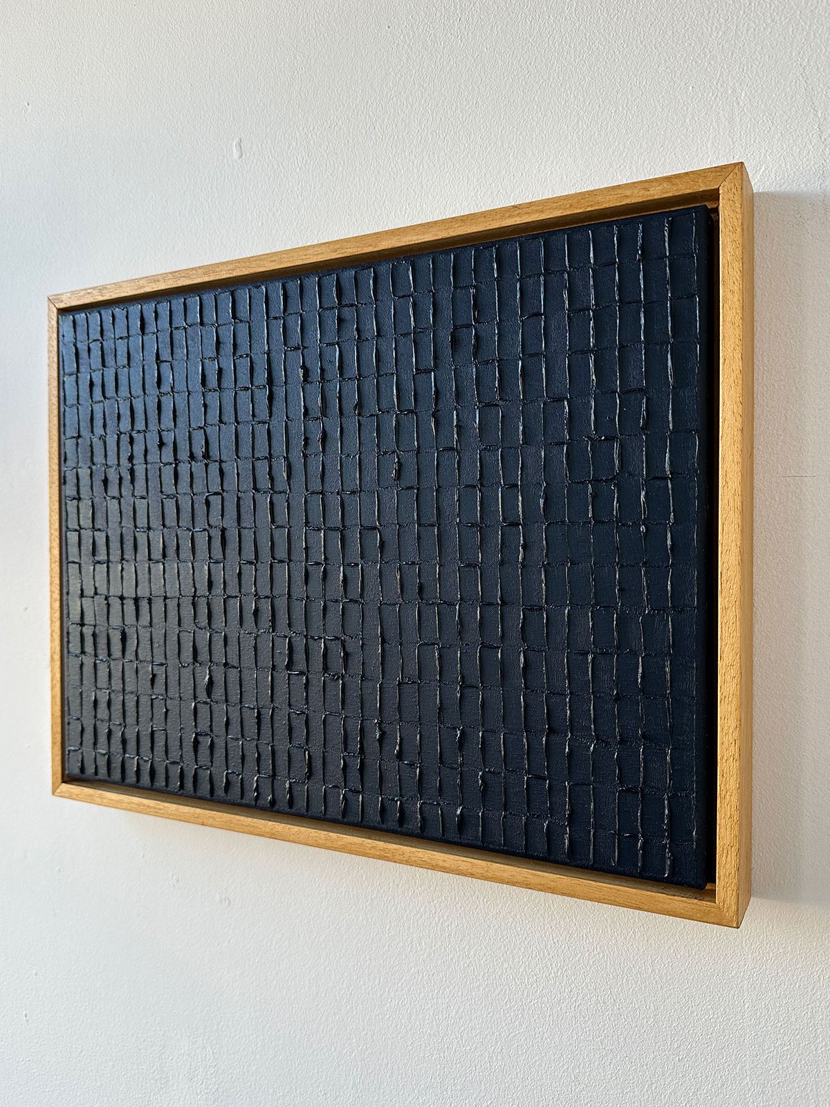 The Prussian Blue artwork by Roi Elmaliah is an oil on canvas rectangular piece with a wooden frame, featuring a textured black surface with raised rectangle grid patterns. Inspired by Amsterdam, it's ready to ship and ideal for display on a light-colored wall.