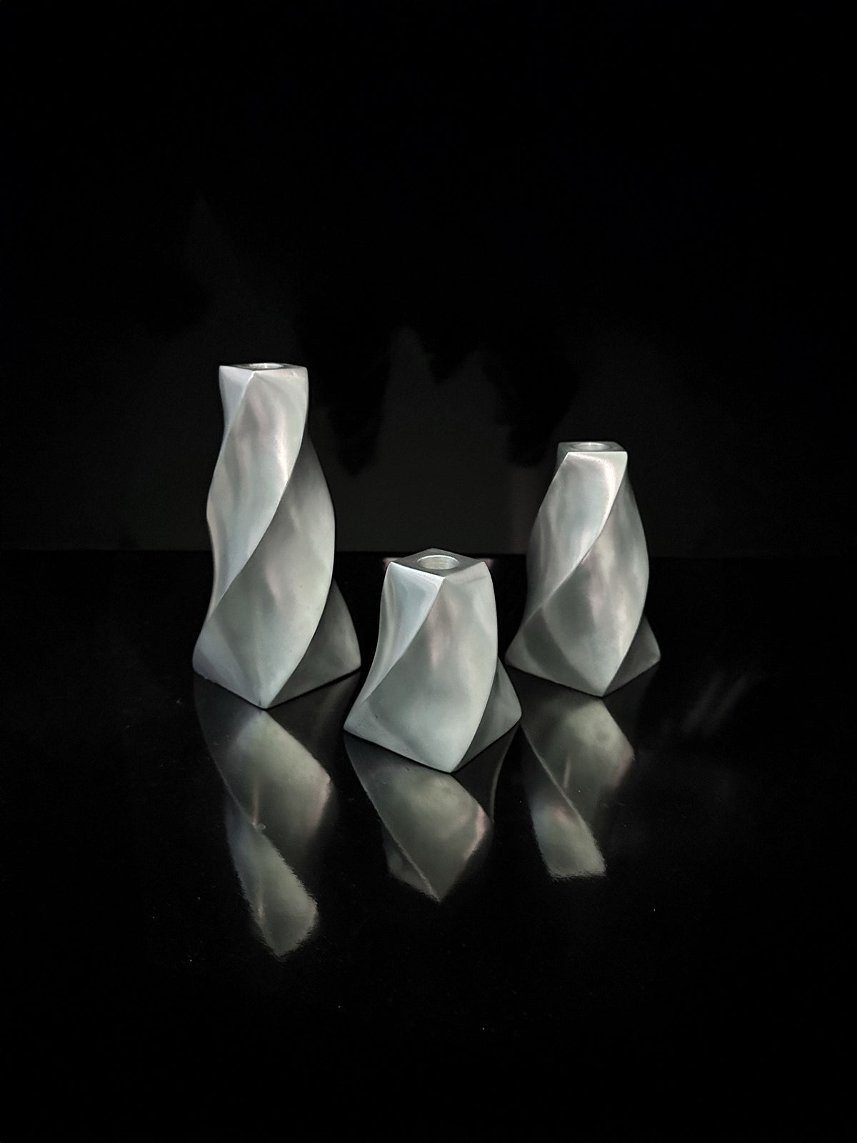 Displayed against a black background are three angular Dodo Vintage candle holders, crafted from aluminium with twisted swirl designs and varying heights. Their metallic surfaces cast delicate reflections on the surface below, evoking an elegant aesthetic.