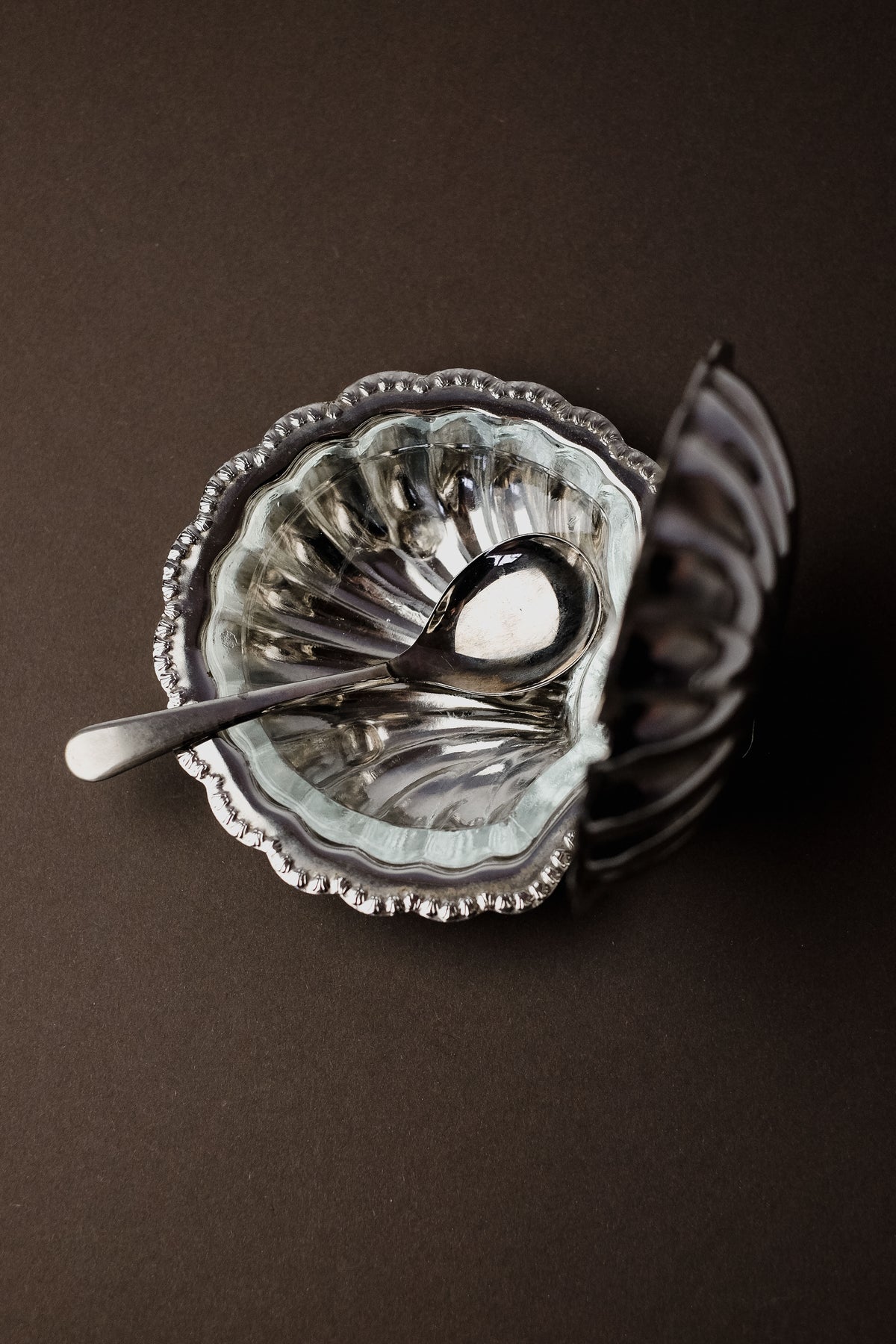 A silver spoon rests in septembre studios' Shell Shaped Butter Dish, showcasing its ornate, scalloped design on a dark surface. The metallic finish and intricate detailing enhance the luxurious dining ambiance, emphasizing rich textures and craftsmanship in dim light.