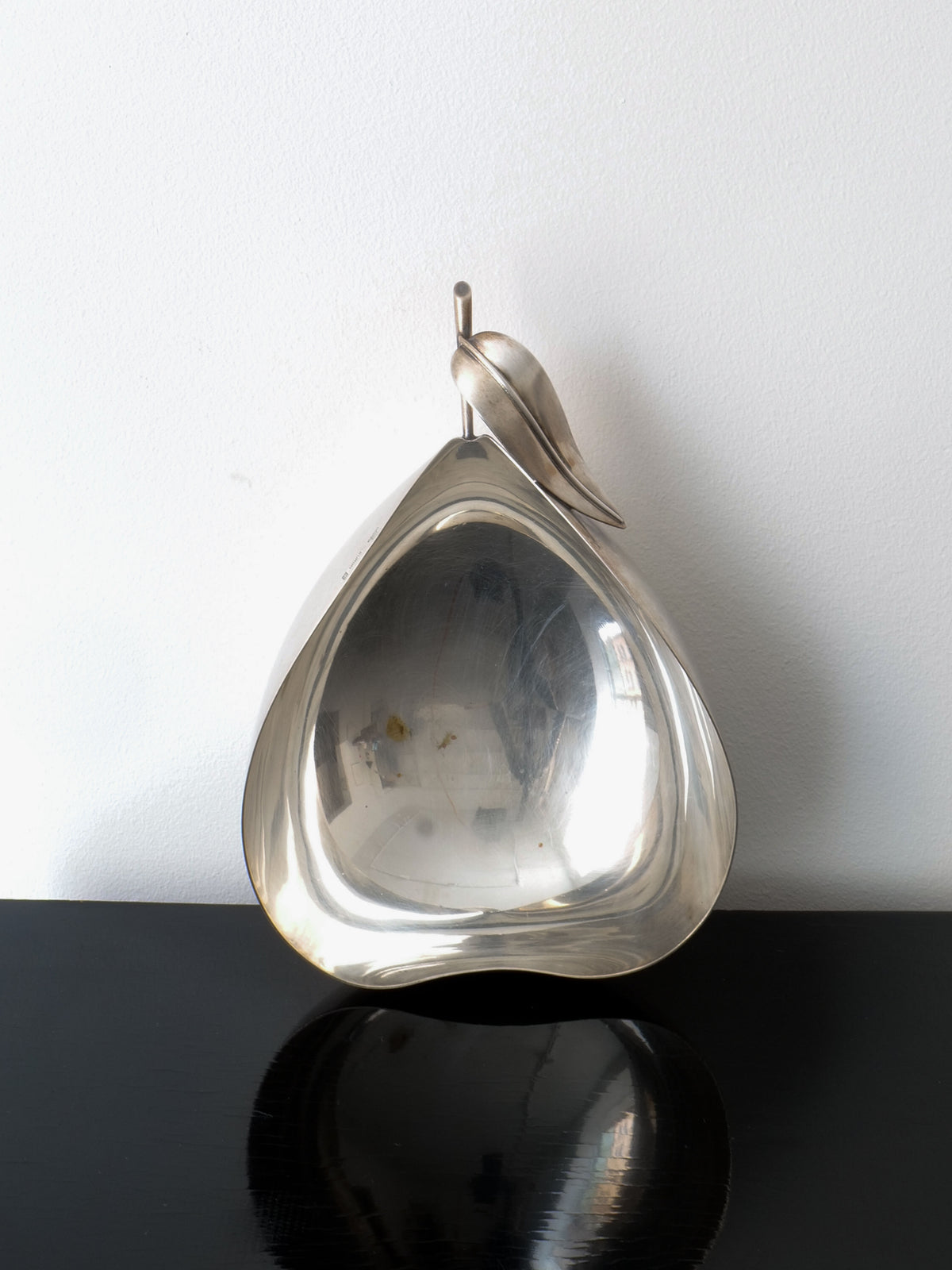 The Christofle Vide Poche Lino Sabattini, from the Collection apart brand, is a silver, pear-shaped bowl featuring a leaf design at the top. It rests elegantly against a white wall on a black surface, with its interior reflecting light beautifully to highlight its smooth finish.
