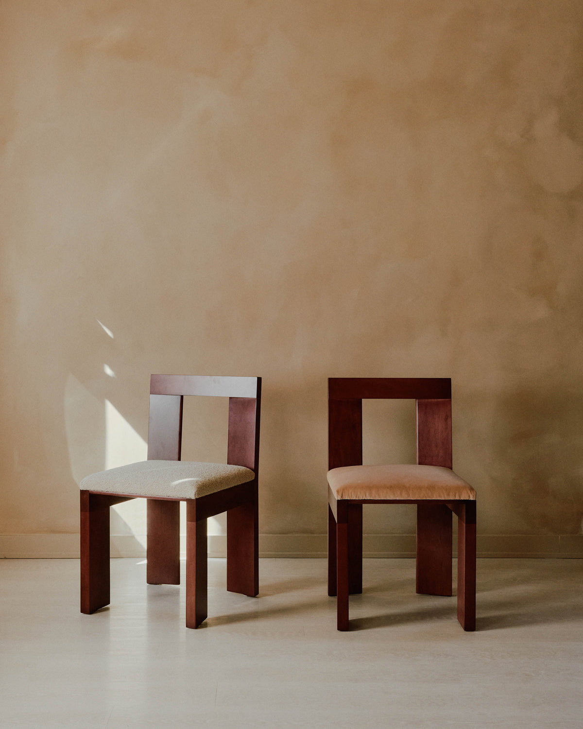 Two Raja Chairs by Juntos Projects are positioned side by side against a beige wall. One chair features a light beige upholstered seat, while the other has a darker beige cushioned seat. The contemporary furniture design and soft lighting lend a calm, serene atmosphere to the scene.