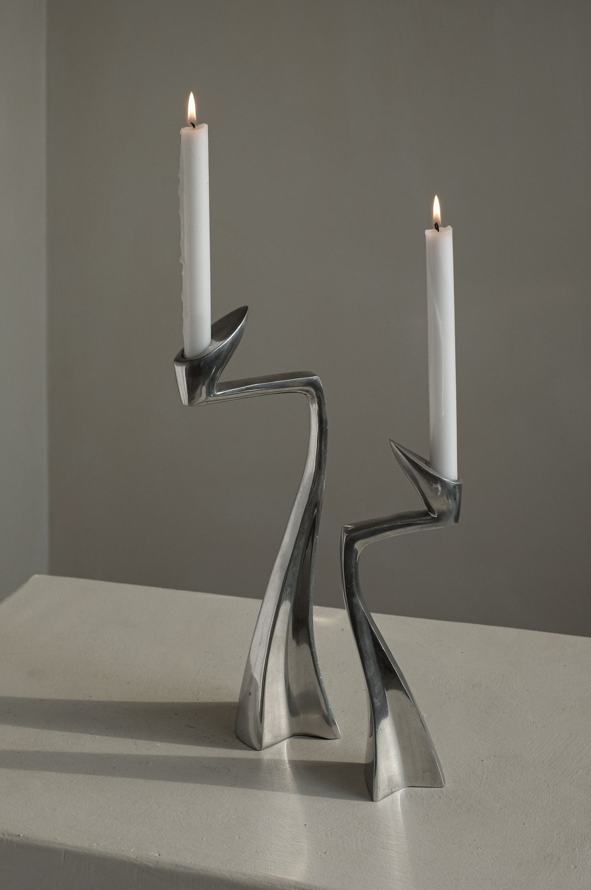 Two white candles burn on the Pair of Candleholders by Matthew Hilton 80's from septembre studios, featuring sleek, twisting designs ideal for design enthusiasts. These collectible pieces sit on a textured white surface with a neutral backdrop.