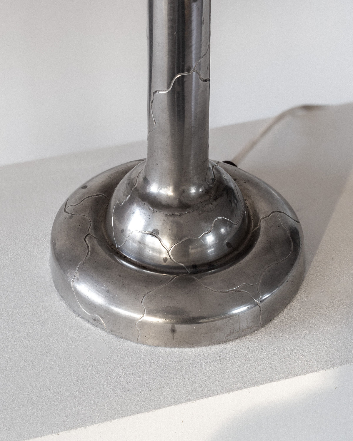 Close-up of a Spigel Art Deco Pewter Lamp base from 1920s Sweden on a white surface. Cracks filled with silvery material mimic kintsugi-style repair, showcasing imperfections. The lamp casts a shadow on the surface.