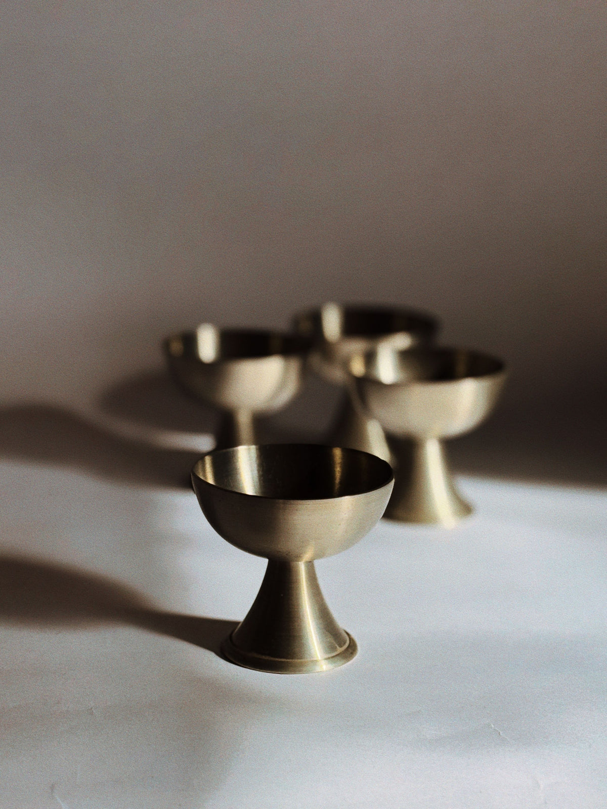 A set of four Huerto brushed metal dessert coupes is arranged on a white surface with soft shadows—three in the background and one in the foreground—all reflecting warm light.