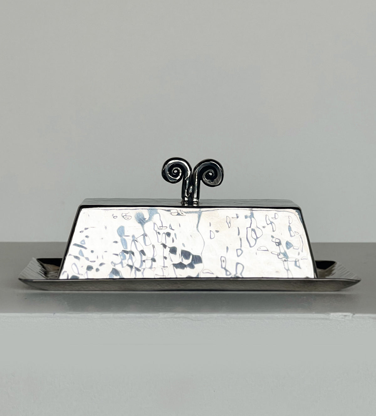 The Spiral Butter Dish by Porter Studio, a handmade silver piece with a hammered texture and decorative handle, shines as it reflects light on a flat surface. Its vintage 1970s charm stands out against the neutral background.