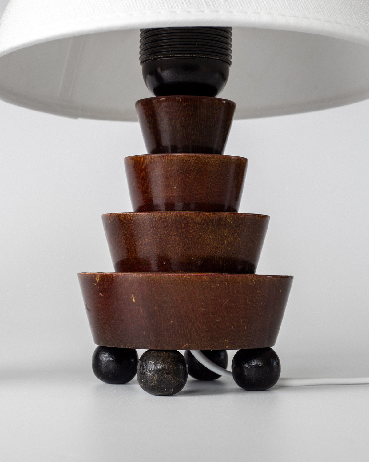 A close-up of the Spigel Art Deco Wooden Table Lamp features stacked round wood segments, four small ball feet, a partially visible white lampshade at the top, and a white power cord.