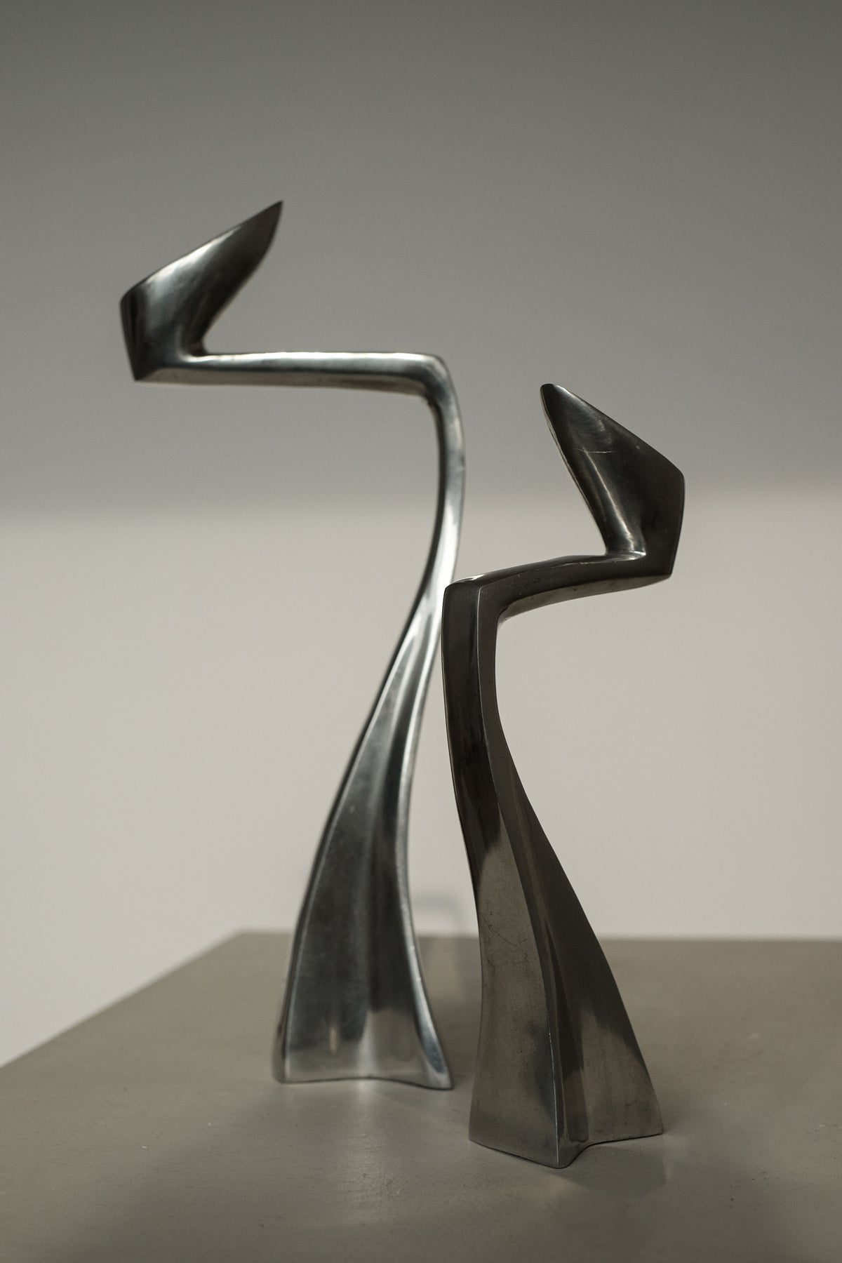 Two abstract, elongated metal sculptures with curved forms resemble the Pair of Candleholders by Matthew Hilton 80's from septembre studios. The taller piece leans slightly over the shorter one, casting captivating shadows and making them coveted collector's items for design enthusiasts.