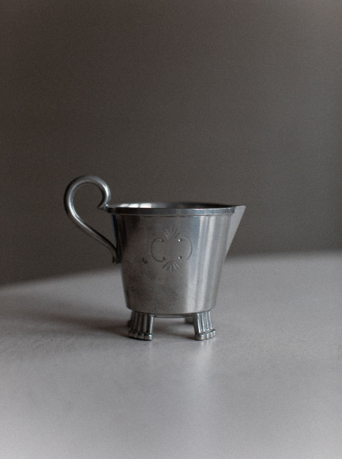 A vintage, silver-colored metal creamer pitcher with an ornate design etched on its body exudes Swedish grace. The Art Deco Swedish Grace Tea-set by Out For Lunch has a small handle and stands on three short, detailed legs, showcasing minimalist charm against a neutral background.