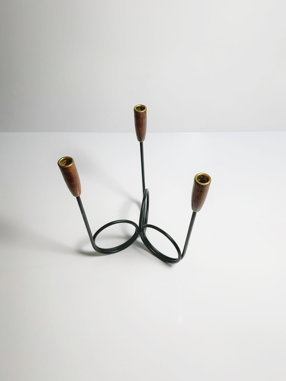 The Danish Candleholder from the 1960s by Dodo Vintage features three metal loops and three vertical candle slots with teak ends, all presented on a smooth white surface. Its modern and elegant design showcases a simple yet stylish aesthetic.