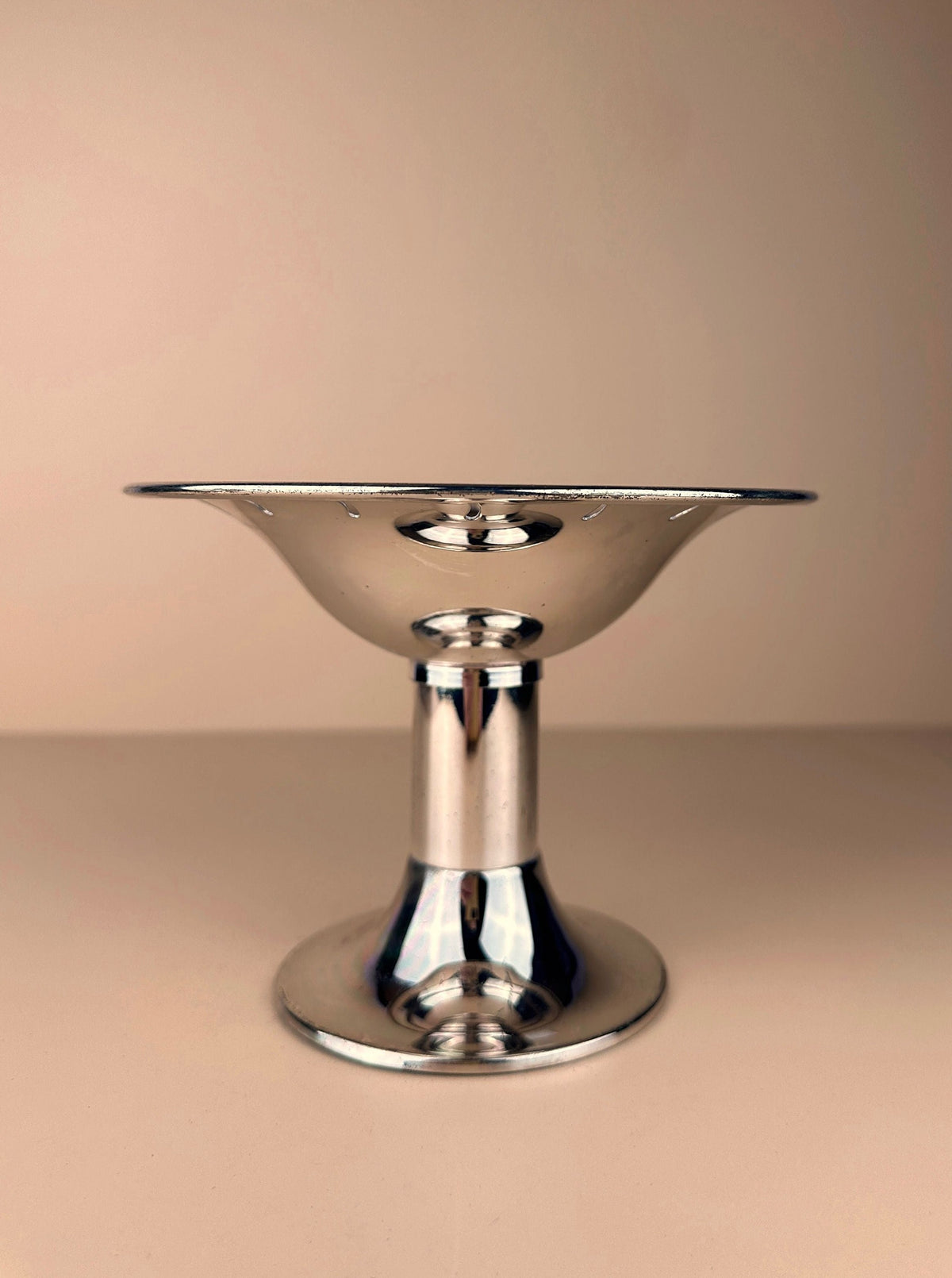 A shiny 1980s silver-plated foot bowl with a wide, shallow circular top and a flared base by Dodo Vintage. The post-modern design stands out against the neutral beige background, highlighting the sleek, reflective surface of this elegant Italian foot bowl.