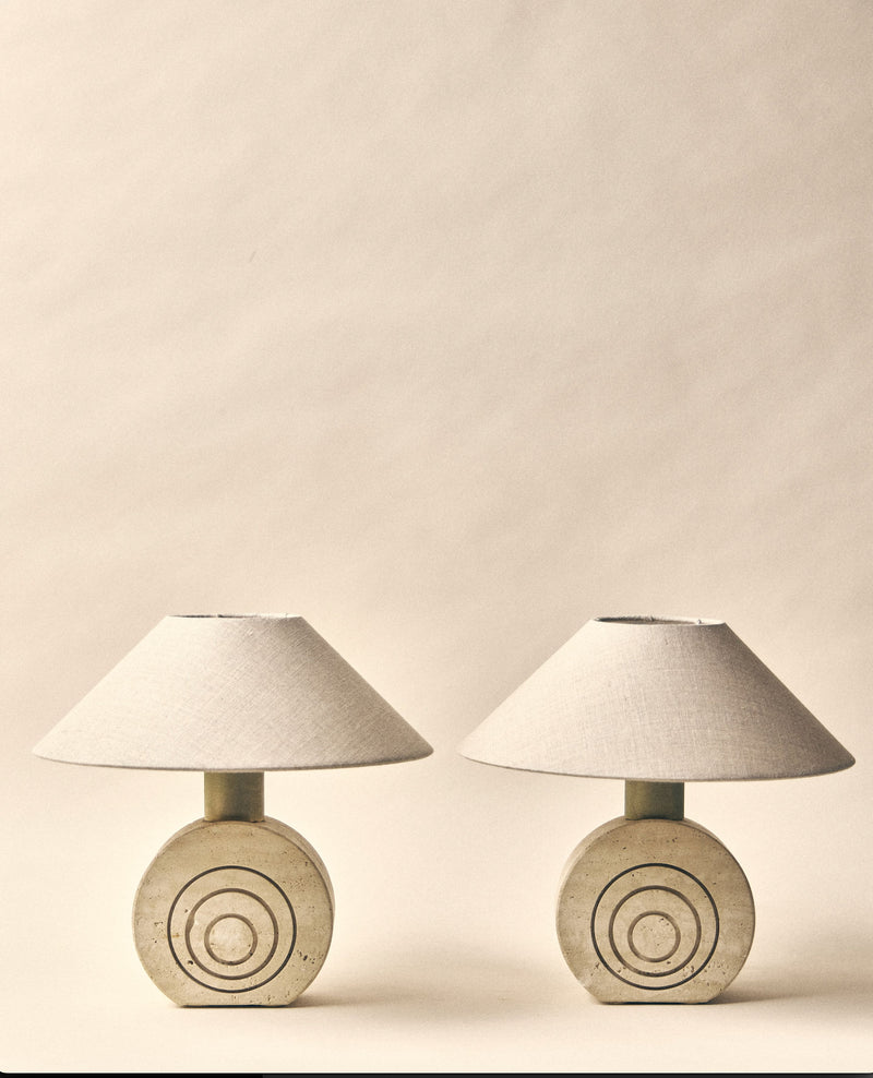 The SÒLER 'Syracuse' Duo Table Lamp features two beige travertine lamps with round, textured bases and concentric circle designs, each topped with a wide, light fabric shade against a neutral background.