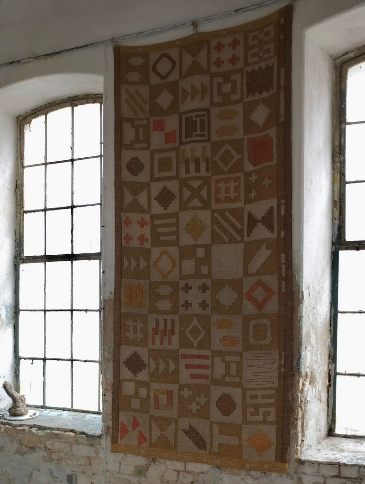A Large Abstract Wool Wall Tapestry by Veter Vintage (Sweden, 1988) hangs between two tall, weathered windows in a rustic room with peeling paint and exposed brick, sunlight streaming in to highlight the tapestry’s vivid patterns.