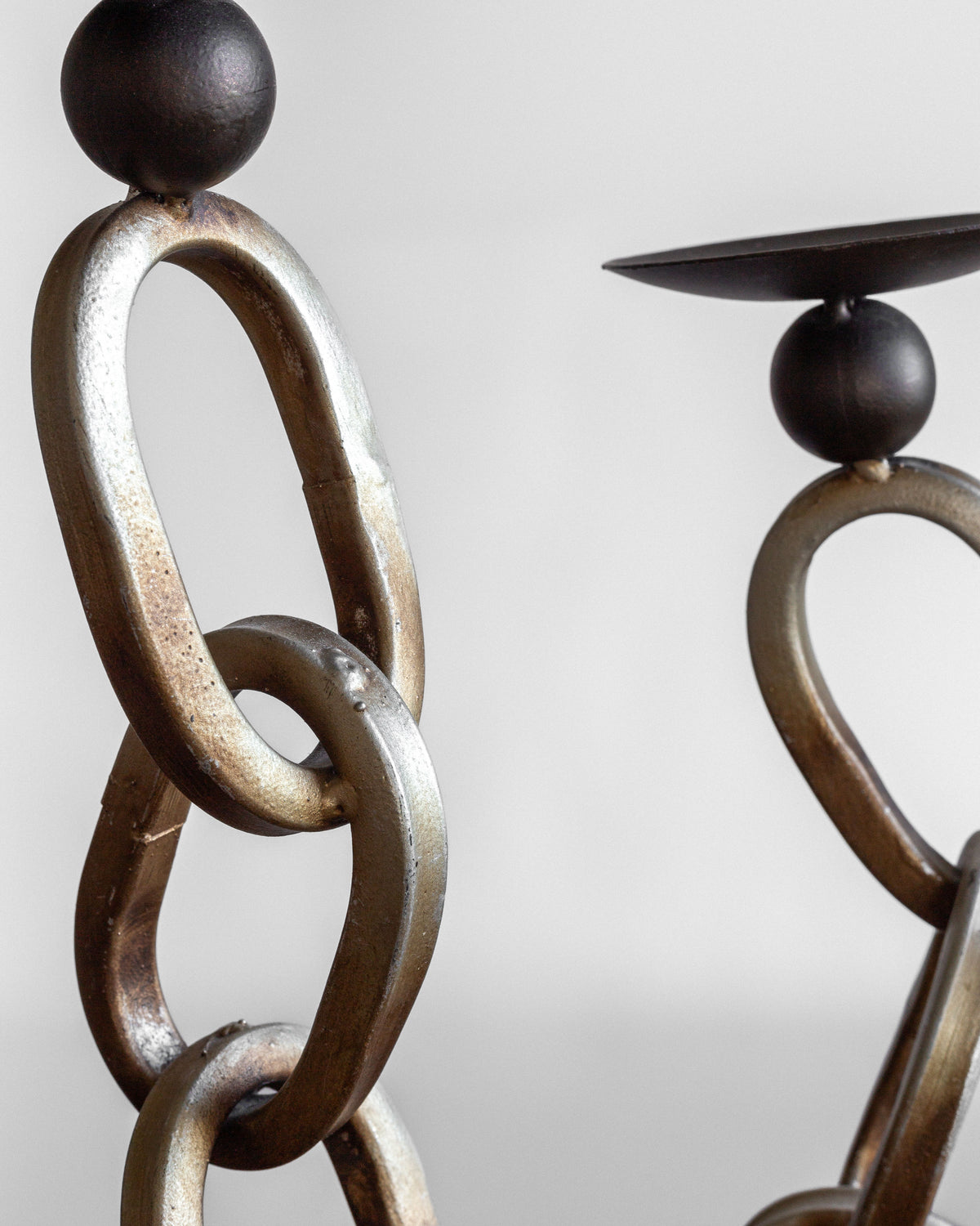 A close-up of the Spigel Postmodern Sculpture Duo showcases large interlocking oval links in a metal chain. The chain culminates in round metal orbs that support small, shallow dishes. This rustic sculpture stands against a plain, light-colored background, highlighting its unique design.