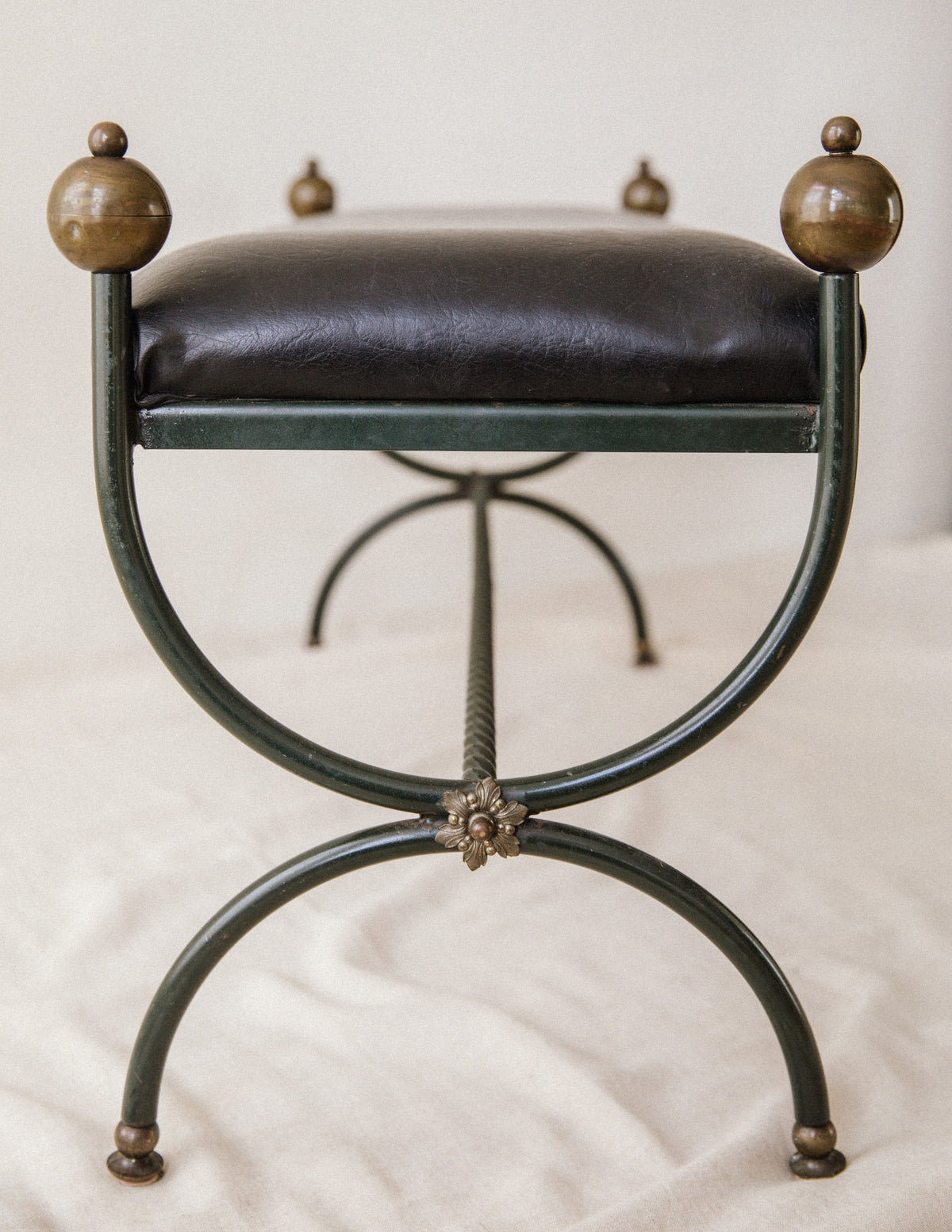 The Arcā Collections Mid-Century Wrought Iron Bench features a black cushioned seat, metal frame with decorative brass knob finials, curved legs, and a floral accent at the joint, shown on a light fabric surface.