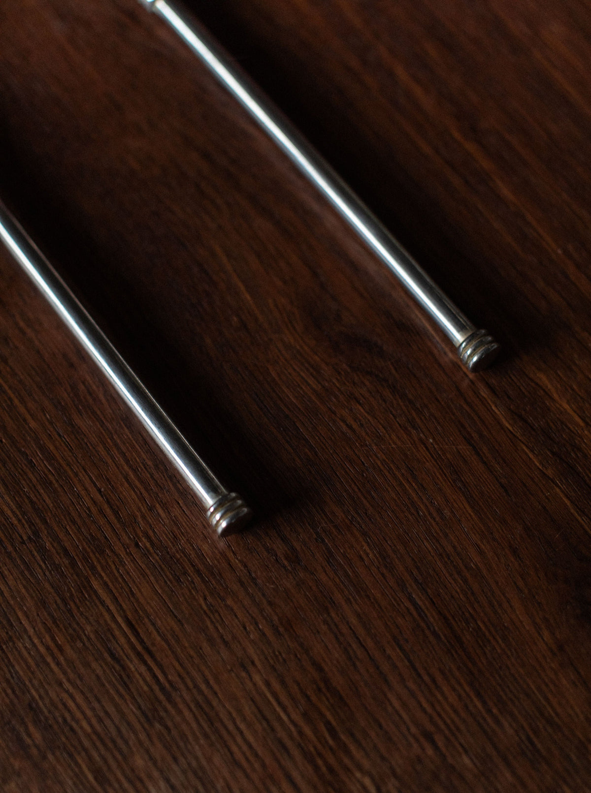 Close-up of two pieces of Art Deco Cutlery from Out For Lunch, featuring long, cylindrical shapes with rounded, decorative ends resting on a dark wooden surface. The focus is on the lower portion of the cutlery, highlighting their smooth texture and metallic sheen against the rich wood grain background, exuding a timeless elegance.