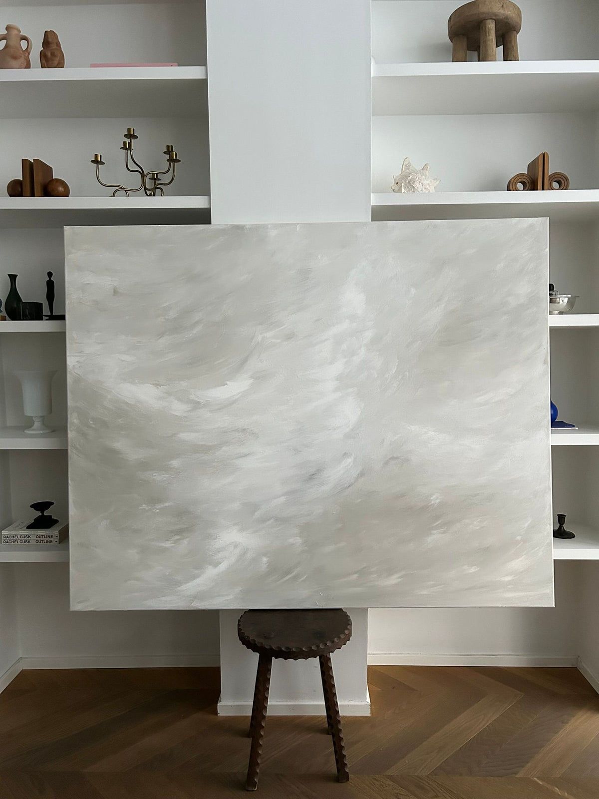 The "Whispering Breeze" painting by Mattias Storm, a large mixed media piece with light grey and white swirls, rests on a wooden stool. It complements white shelves decorated with sculptures, vases, and a candelabra above a classic herringbone floor.