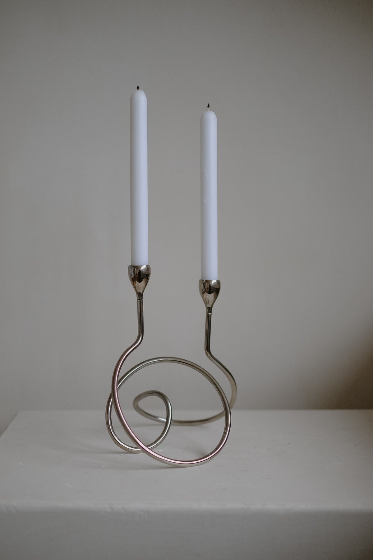 The septembre studios "Loop" Candle Holder showcases a minimalist chrome design with swirling loops and features two unlit white dinner candles, set against a neutral background.