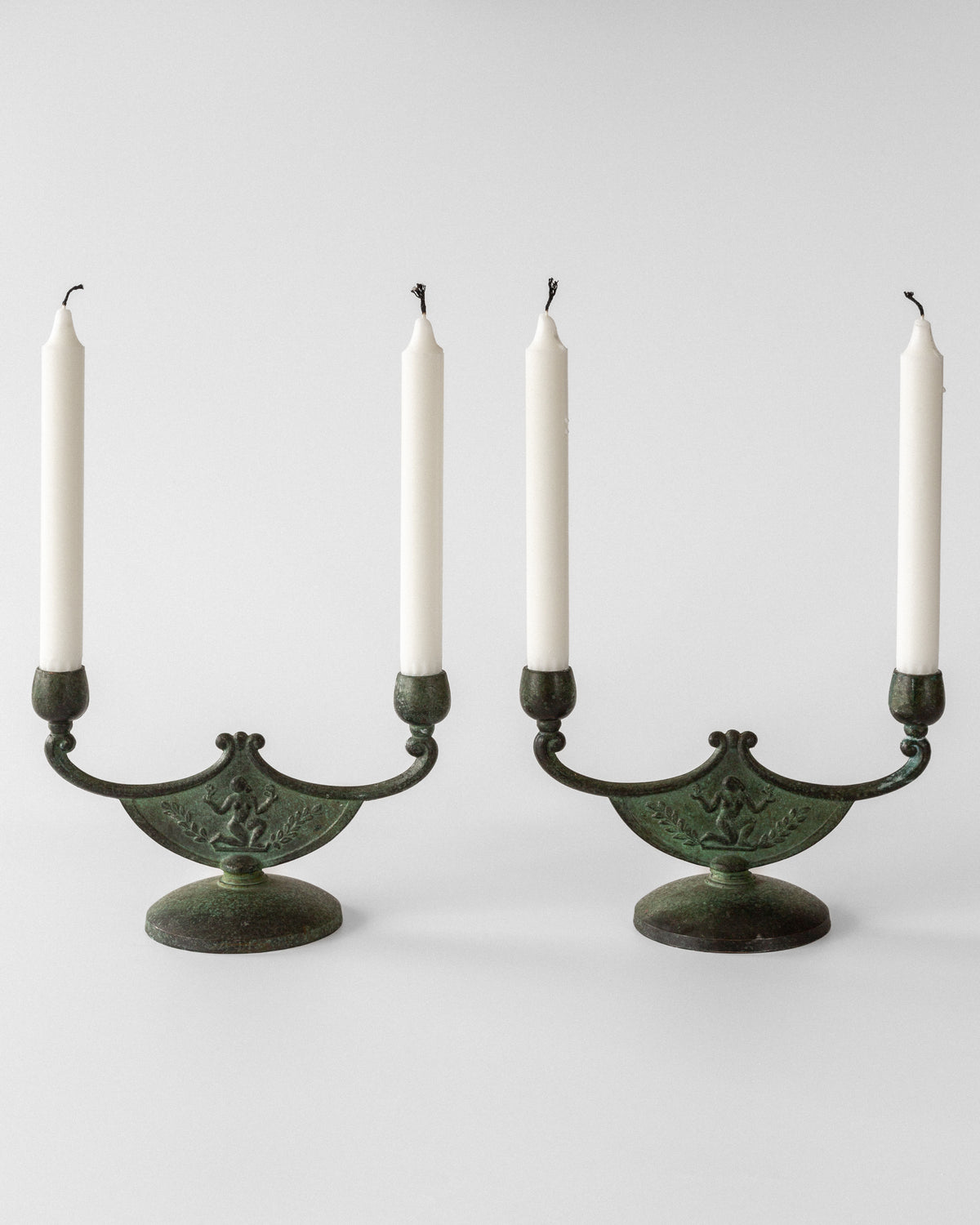 The Spigel Pair of Art Deco Bronze Candleholders exhibit exquisite Swedish metalwork with intricate designs and rounded bases, each holding a tall white candle, set against a plain white backdrop.