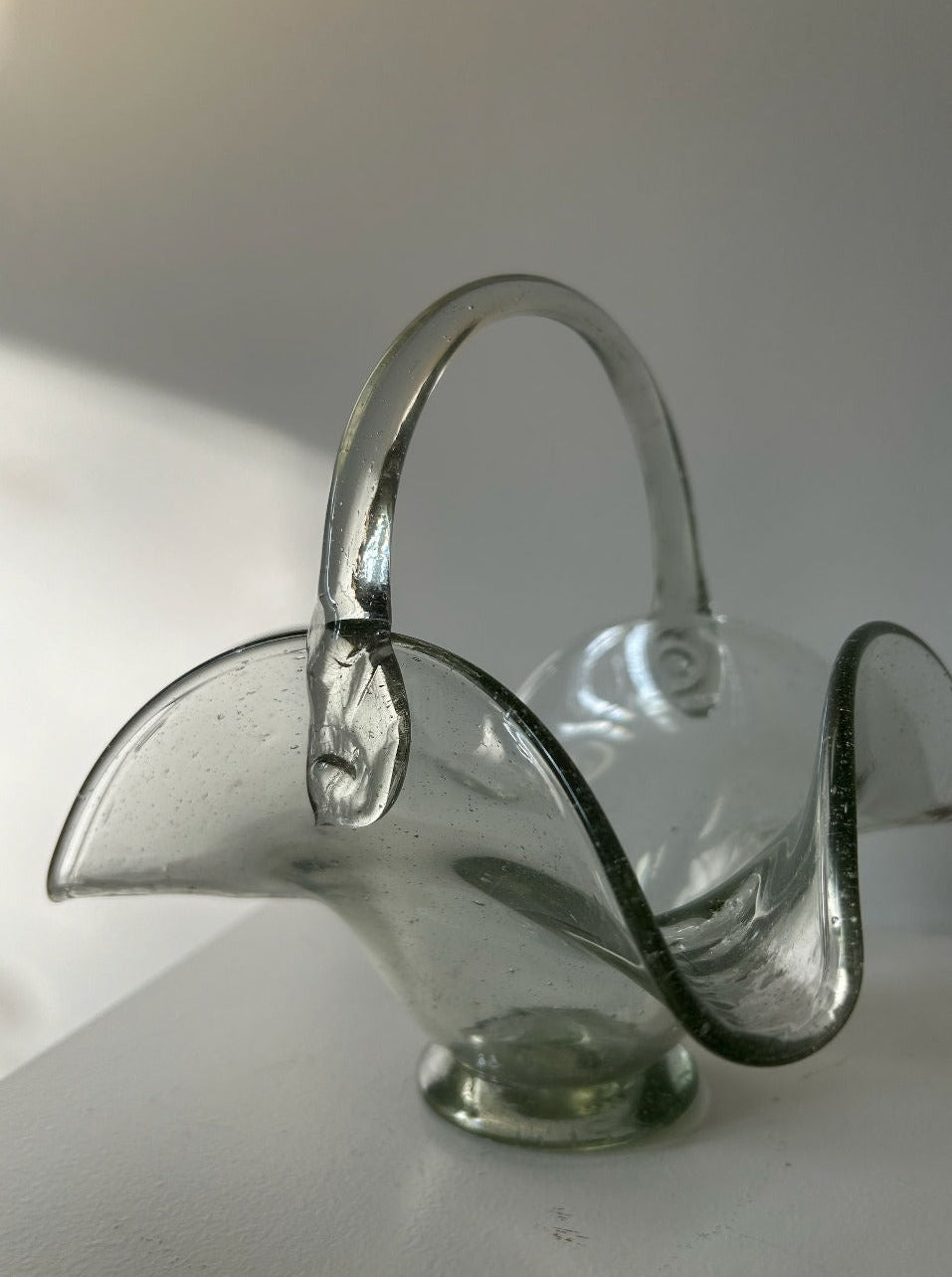 A delicate, transparent glass basket with a curved handle and wavy edges sits on a white surface. Crafted from thick quality glass reminiscent of Les Objets Amsterdam's 70's Vintage Glass Basket, it features slight imperfections that add charm to the piece. Soft light casts gentle shadows, highlighting the basket's elegant shape.