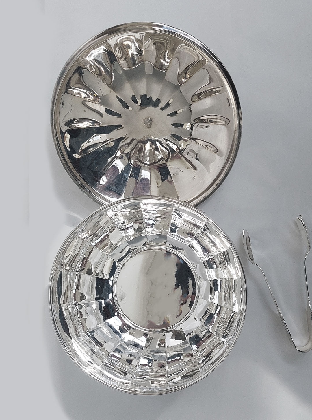 The image captures a refined tableau featuring two polished, reflective Les Objoies Large Sugar Bowls—one placed face-up and the other face-down. To the right, a pair of serving tongs complements the setup. The bowls, resembling silver-plated sugar bowls, showcase a ribbed design with curved indentations along their sides.