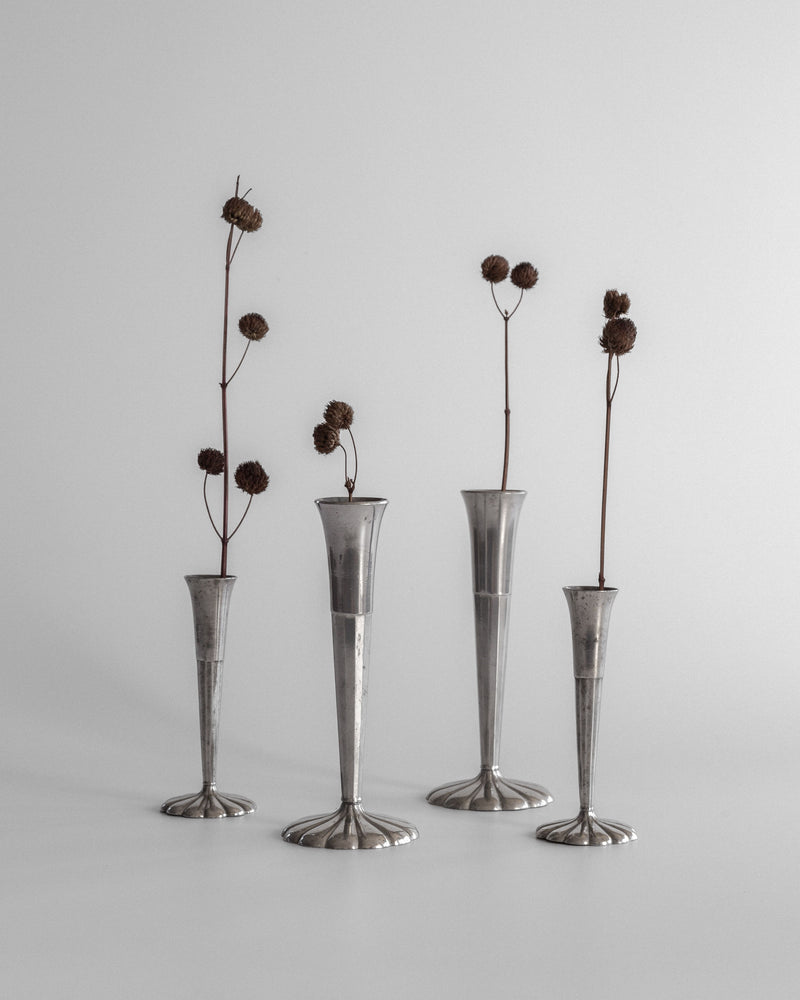 The Spigel Set of Four Art Deco Metal Vases, each tall and slender with varying heights, feature single dried flower stems with round seed heads. This elegant arrangement on a plain surface against a neutral backdrop embodies the timeless essence of Scandinavian design.