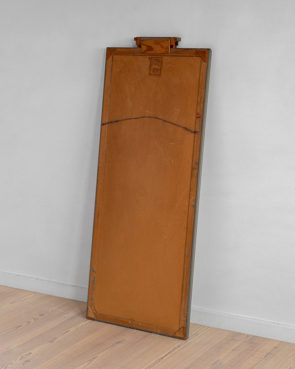 A rectangular, wood-framed Art Deco piece by Spigel, reminiscent of 1930s Sweden, adorns a light gray wall. The back reveals a brown backing and hanging wire. This mirror by J.A. Edenholm leans gracefully against the light wooden floor.