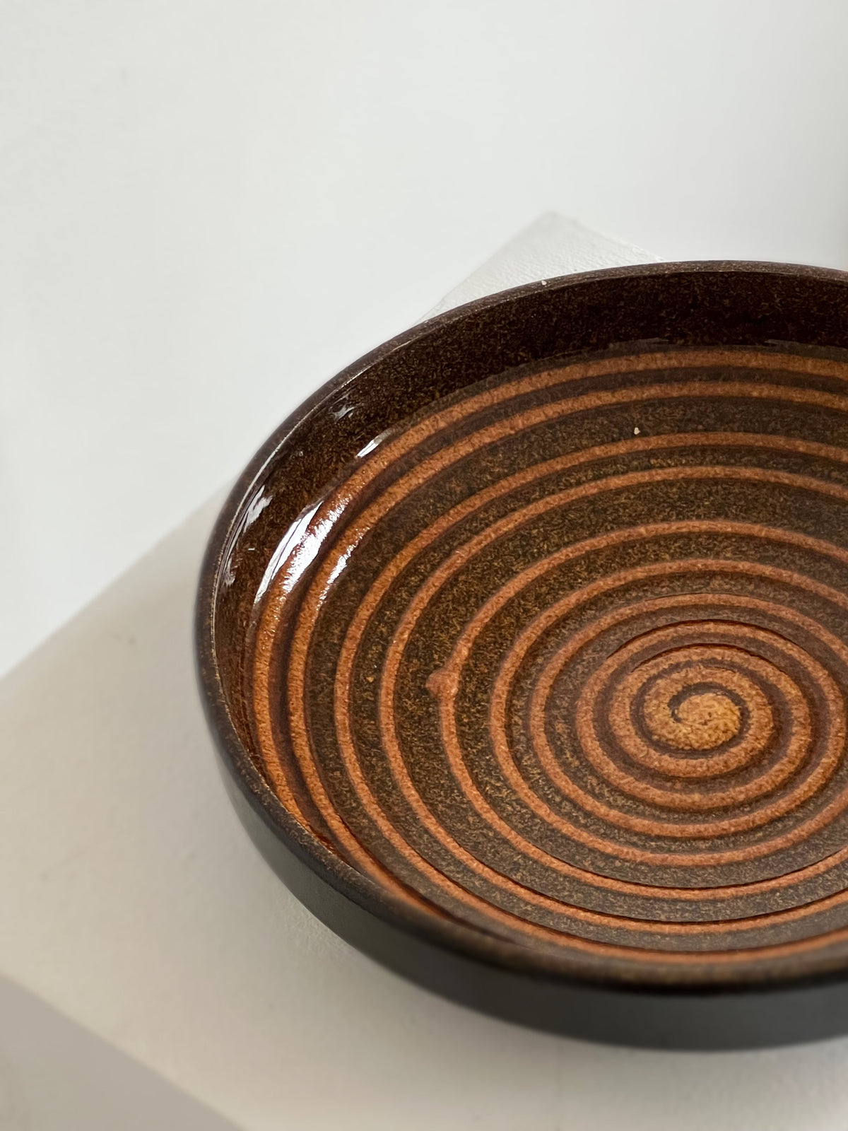The Porter Studio Spiral Ceramic Dish features glossy concentric brown and tan circles on a neutral background, creating a captivating, hypnotic swirl pattern perfect for kitchen use.