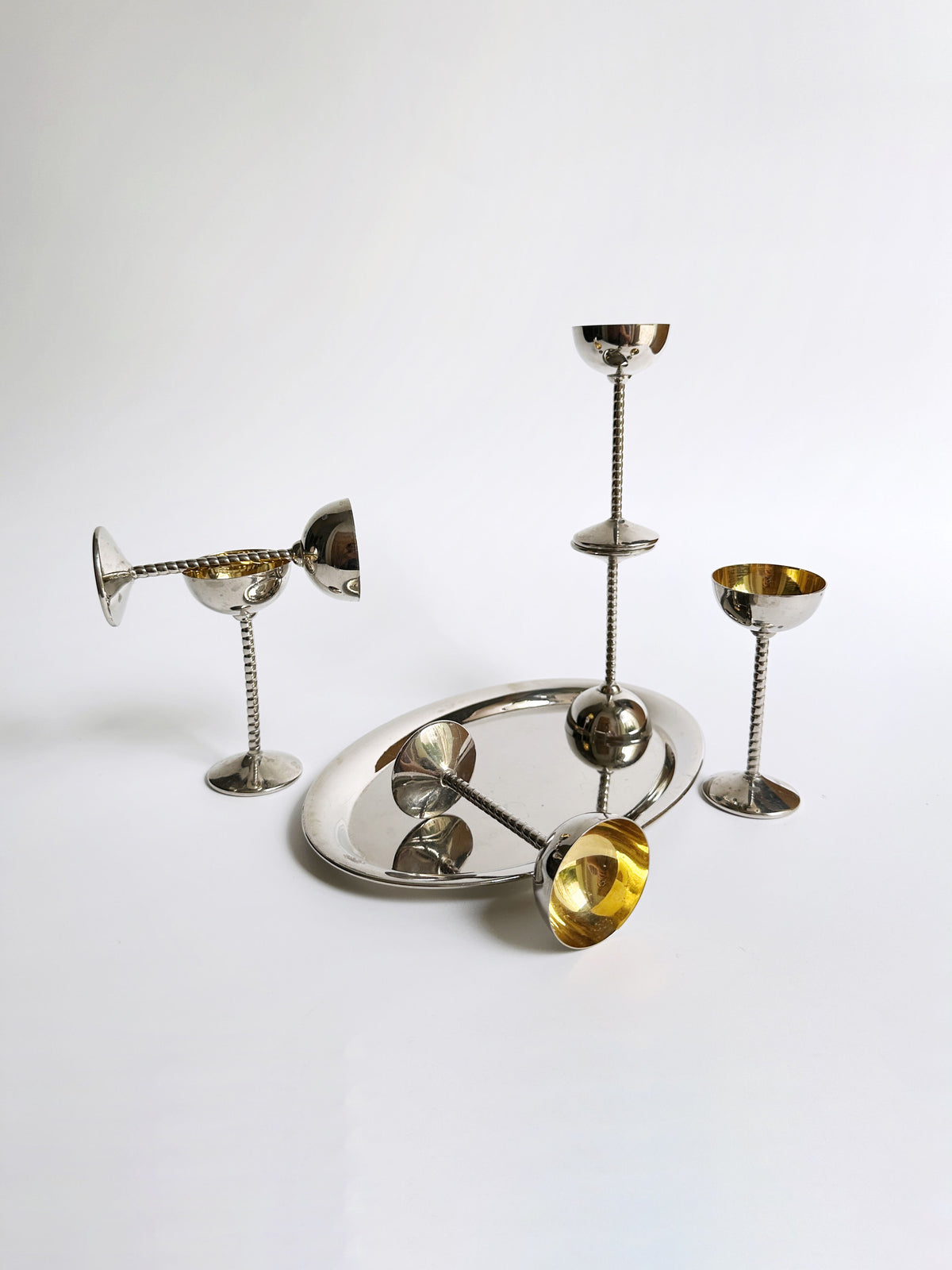 A collection of Art Nouveau liqueur goblets from Dodo Vintage, featuring twisted stems and golden interiors, is artfully arranged on a reflective silver tray. One goblet is tilted, while two others stand upright on the white surface, evoking an elegant display. The background remains a pristine white.