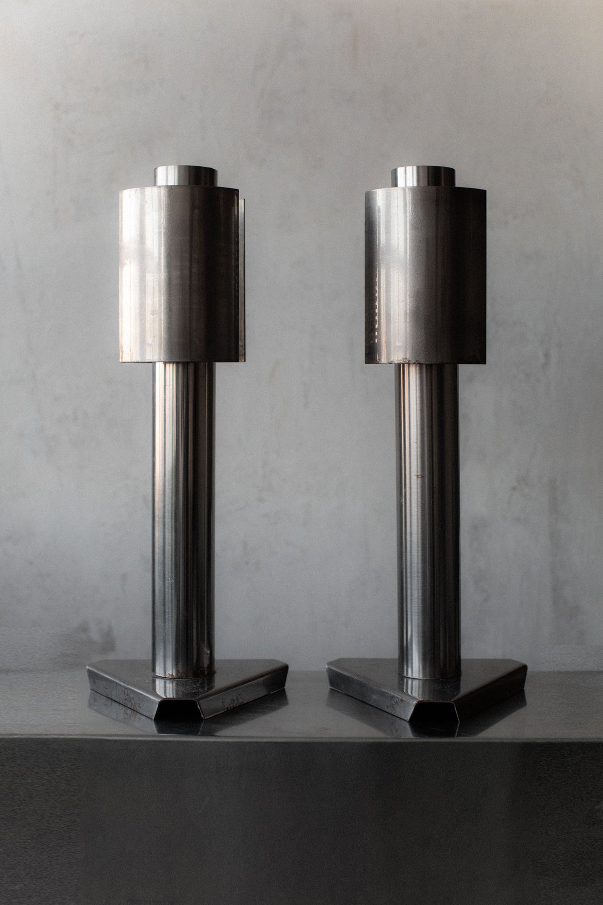 Two tall, cylindrical sculptures stand side by side on a flat surface. Each has a geometric, industrial design with a vertical orientation and a hexagonal base, exuding the 1960's Space Age aesthetic of Out For Lunch lamps against the plain gray background.