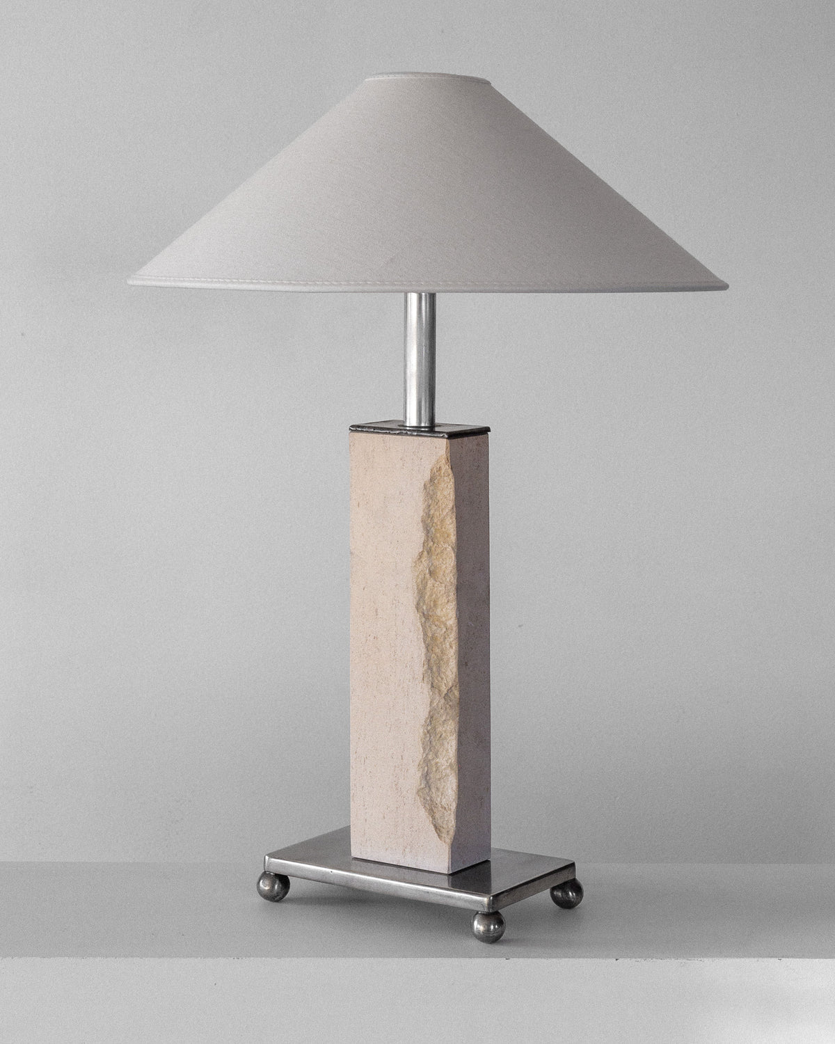The Spigel Modernist Travertine and Metal Table Lamp features a mid-century modern design with a rough textured rectangular stone base on a metal platform with round feet. It has a large light gray conical fabric shade against a plain light-colored background.