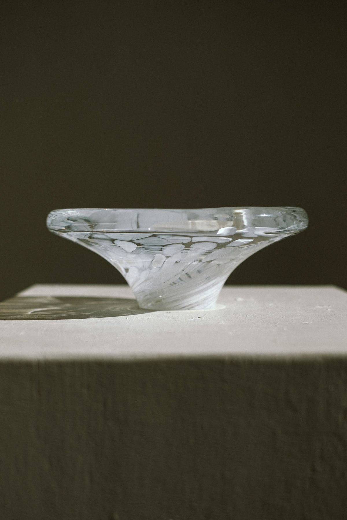 The White Bowl Tortoise Glass by septembre studios, featuring a sculptural swirling design, is displayed on a textured white pedestal against a softly lit, plain background.