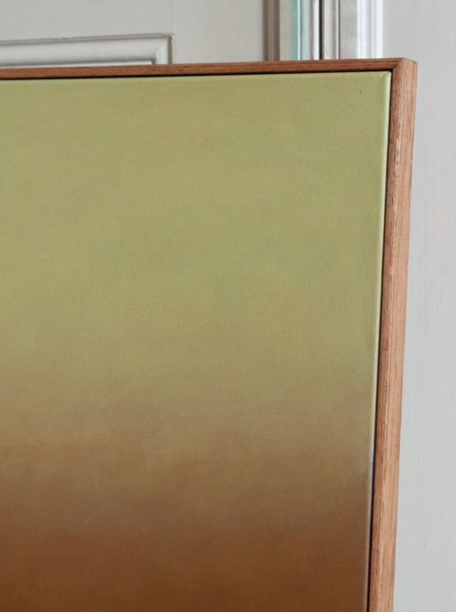 A close-up photograph of a corner of an acrylic on canvas painting, titled "Something(s) in the air, 2024" by Rebecca von Matérn, with a smooth gradient transitioning from light green at the top to brown at the bottom, framed by a thin, light oak frame. The painting is propped against a white paneled wall.