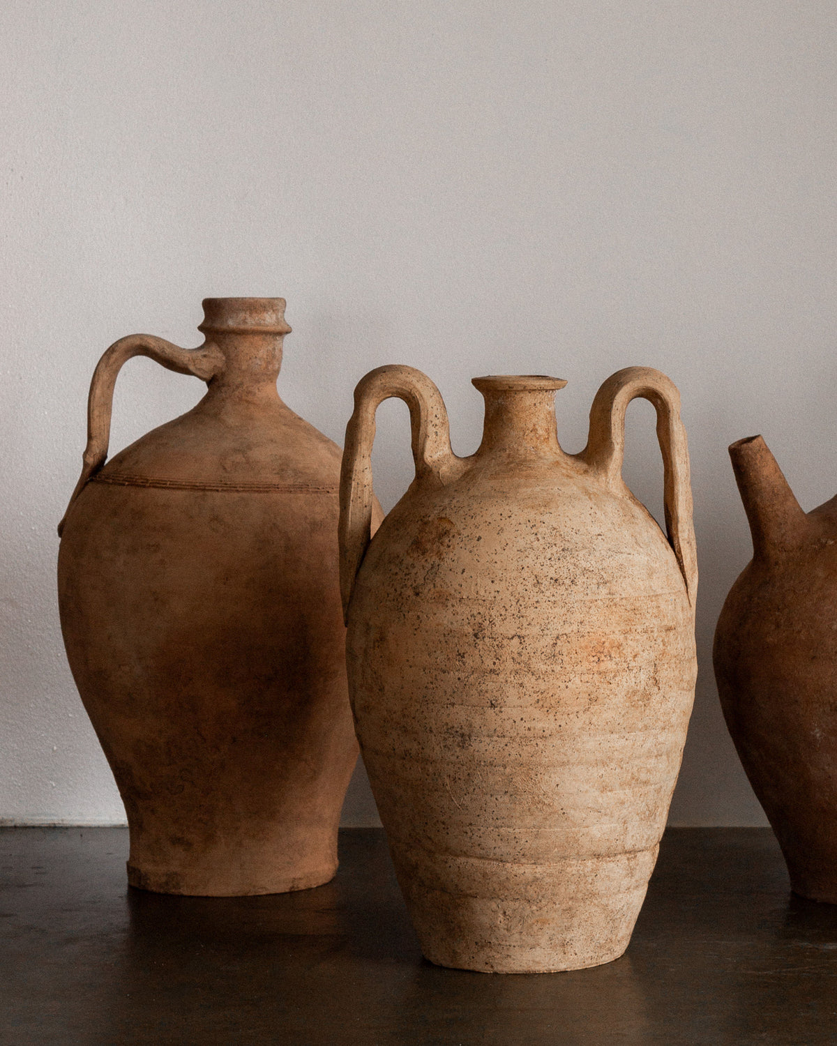 Three hand-crafted Early 20th Century European Olive Jars by Spigel are displayed against a plain background. Each jar has two handles and a narrow neck, exuding a rustic, historical aesthetic with lighting that highlights their earthy tones and timeworn appearance.
