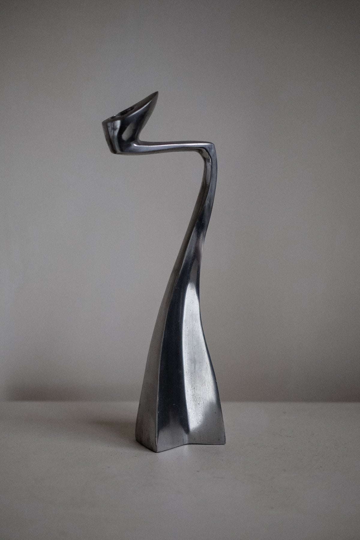 A sleek, abstract metallic sculpture with a curved form stands against a plain background. Reminiscent of the Pair of Candleholders by Matthew Hilton 80's from septembre studios, it features a twisted design with a pointed tip at the top, evoking modern art's essence.