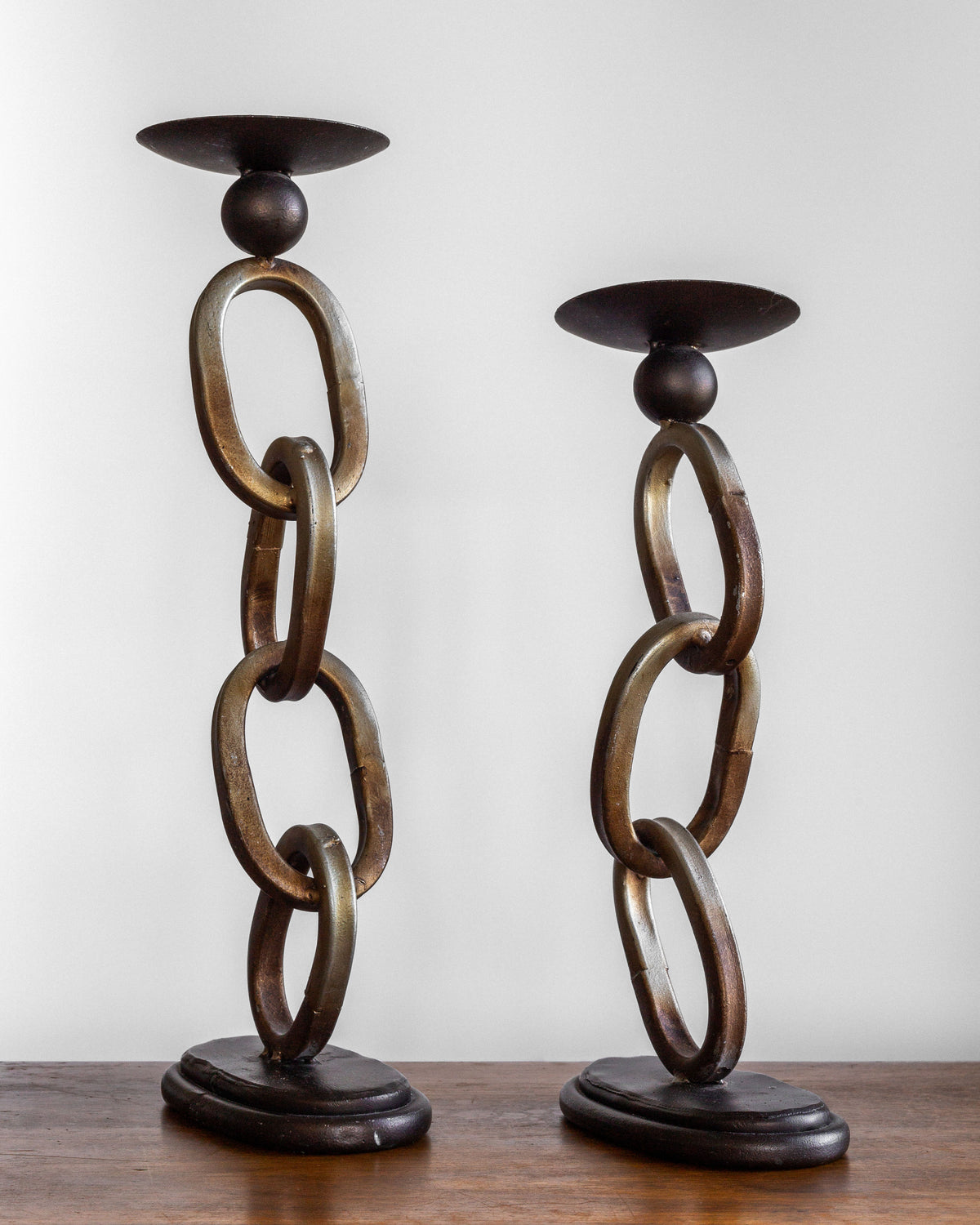 The Spigel Postmodern Sculpture Duo features two modern, artistic candle holders shaped like vertical chains with large, oval metal links. One is slightly taller than the other. With a rustic appearance, they both rest on a dark wooden base and are placed on a wooden surface against a plain white background.