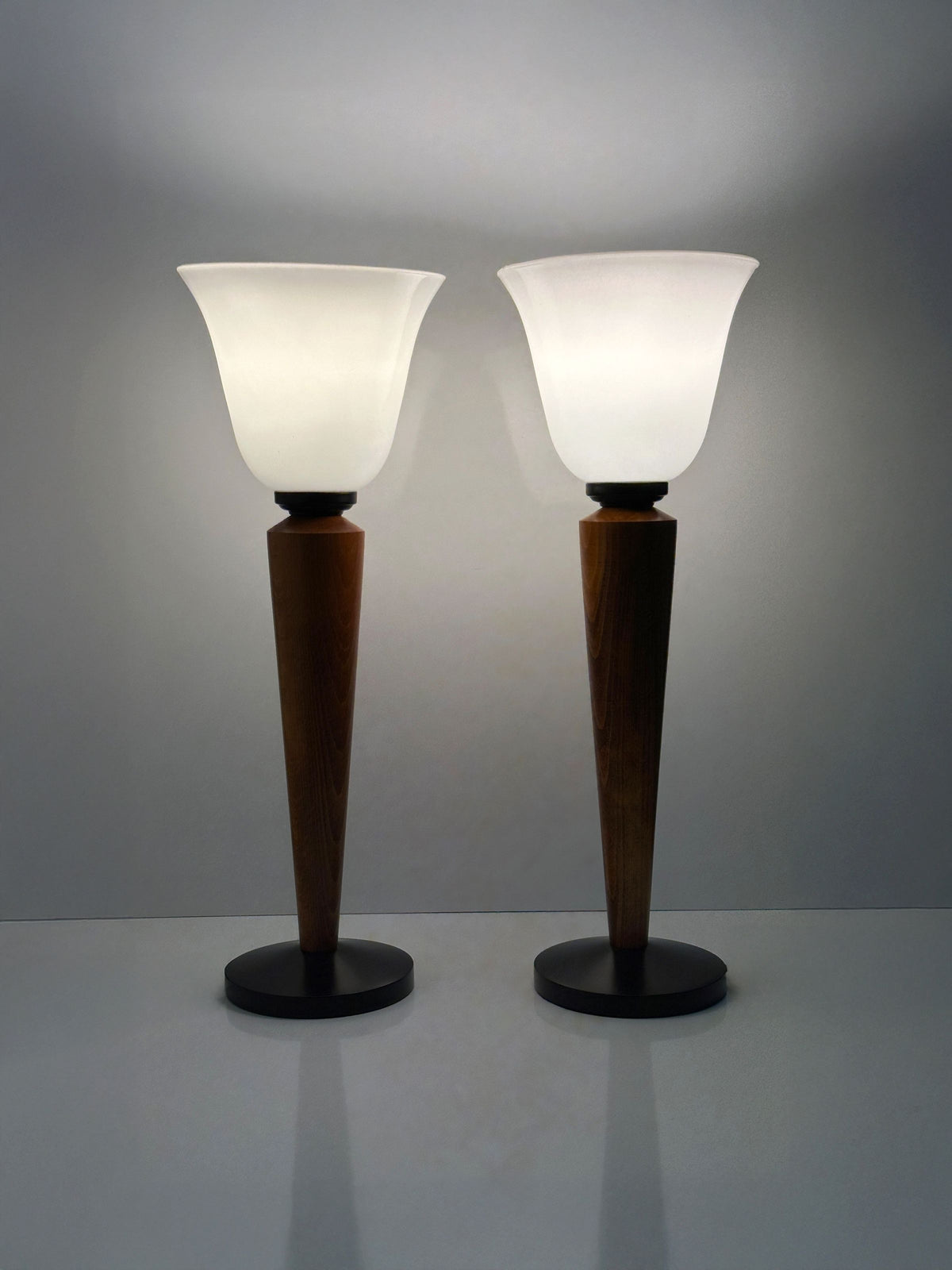 The Pair of Art Deco Chalices with Teak Wood by Dodo Vintage stand side by side on a smooth surface against a plain background, featuring frosted glass shades and teak stems. They emit a soft, white glow, casting gentle reflections below.