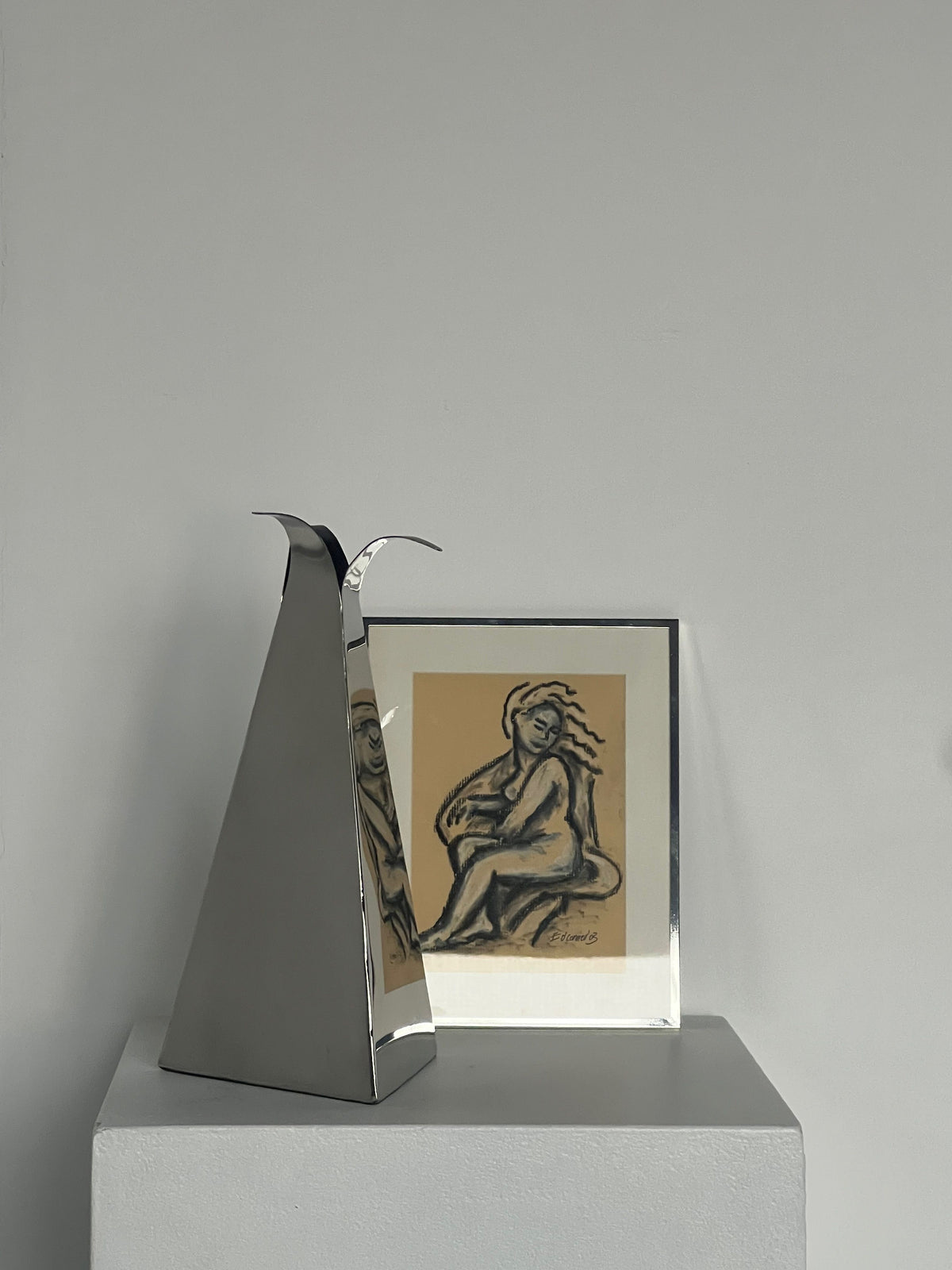 A tall, triangular silver vase with sleek curves sits on a white pedestal. Next to it is "Pastel Drawing by E.D Connor" in a polished chrome frame by Porter Studio, resembling a life drawing of a seated figure. The plain background enhances the minimalist and contemporary display.