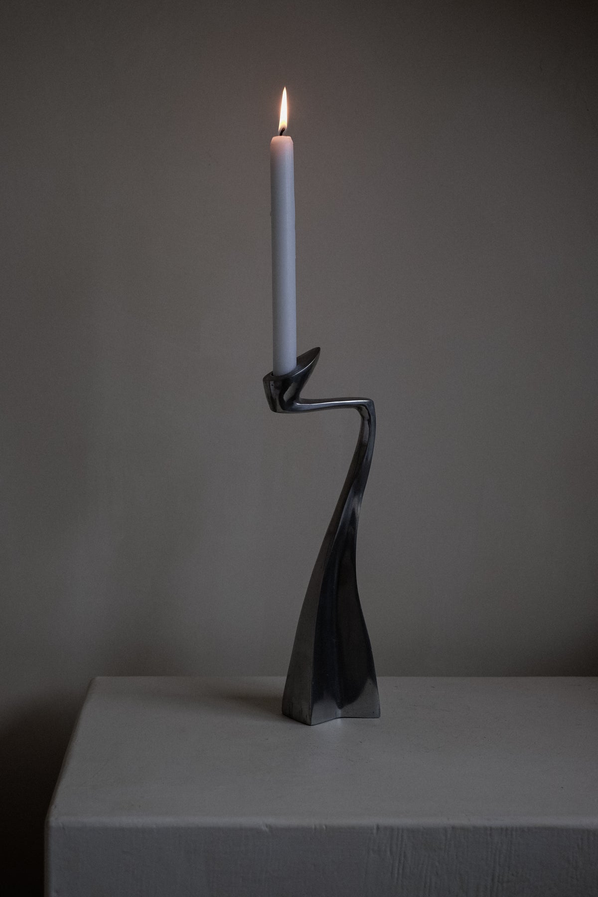 A Pair of Candleholders by Matthew Hilton 80's from septembre studios graces a smooth, neutral surface. The modern, twisted design and candlelight softly illuminate the minimal background, casting subtle shadows—a must-have for design enthusiasts and collectors alike.