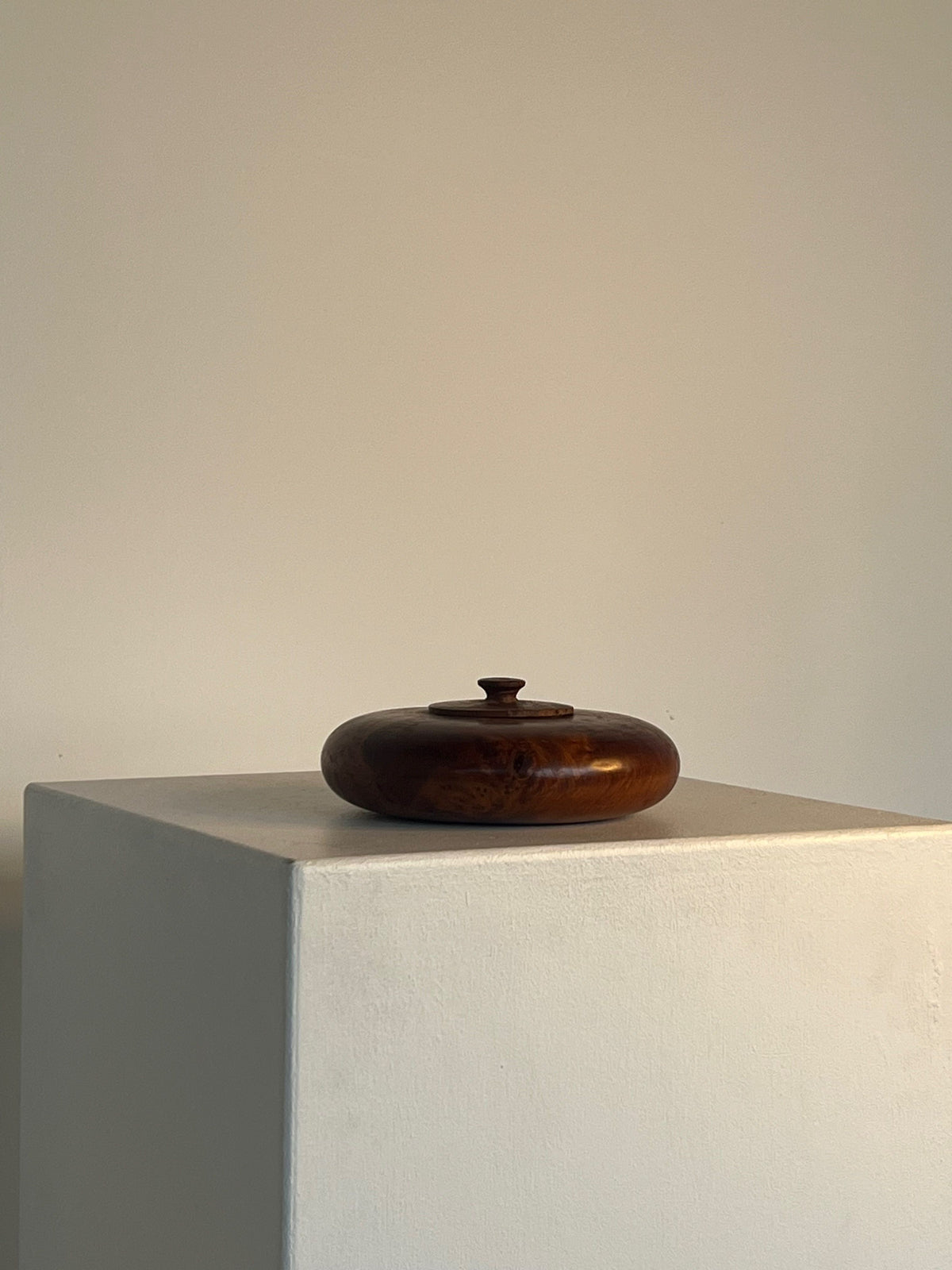 A round and smooth wooden creation from Porter Studio, the Walnut Burl Shallow Bowl With Lid, rests elegantly on a minimalist white pedestal against a plain, light-colored wall. The simple yet sophisticated setting accentuates the exquisite craftsmanship of this remarkable piece.