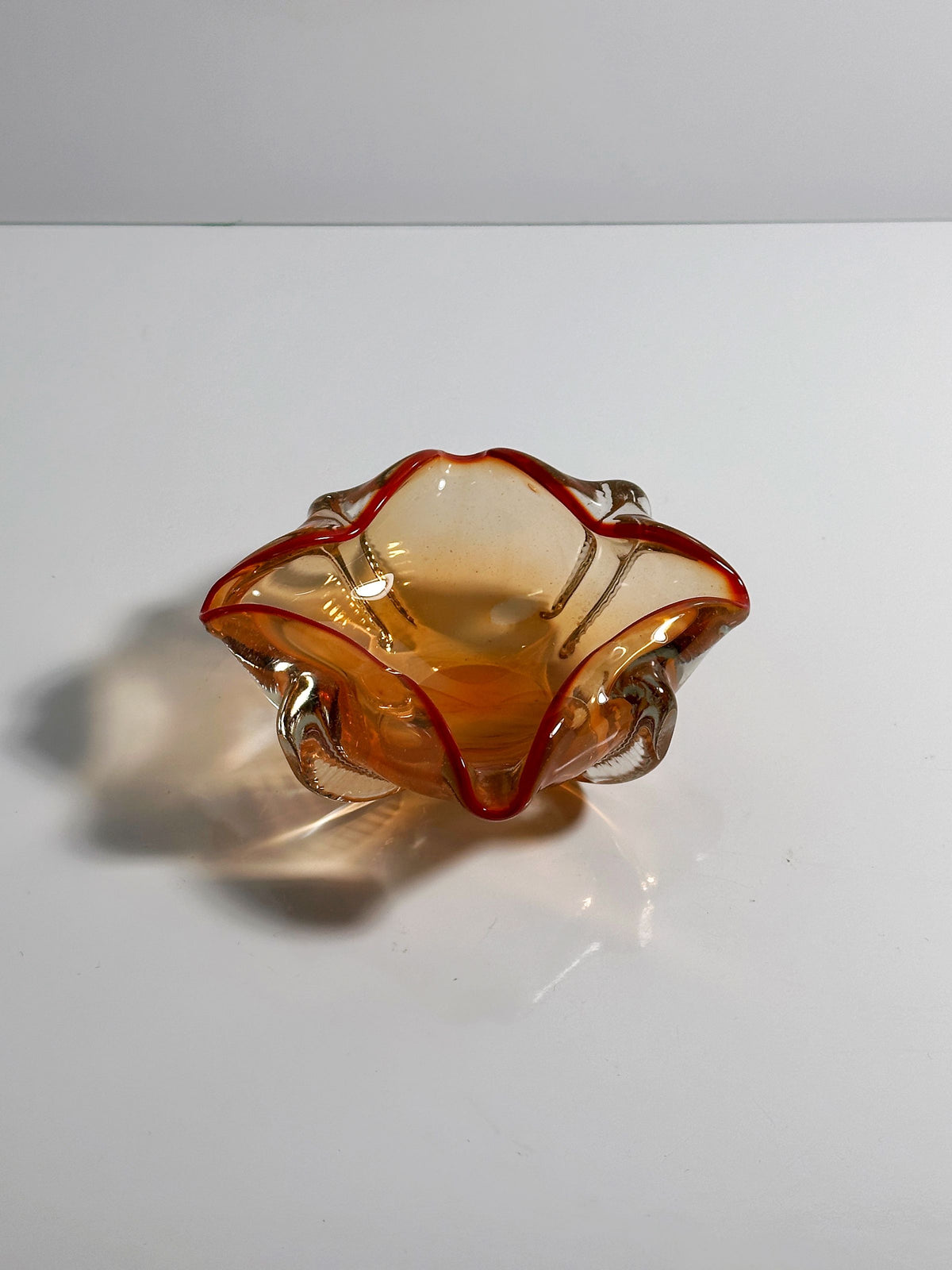The Venetian Ashtray by Seguso Murano from the 1950s, offered by Dodo Vintage, is a small, coral-colored glass piece with wavy scalloped edges and red accents along the rim. Its elegant nautical design casts shadows on the white surface beneath it, adding depth.